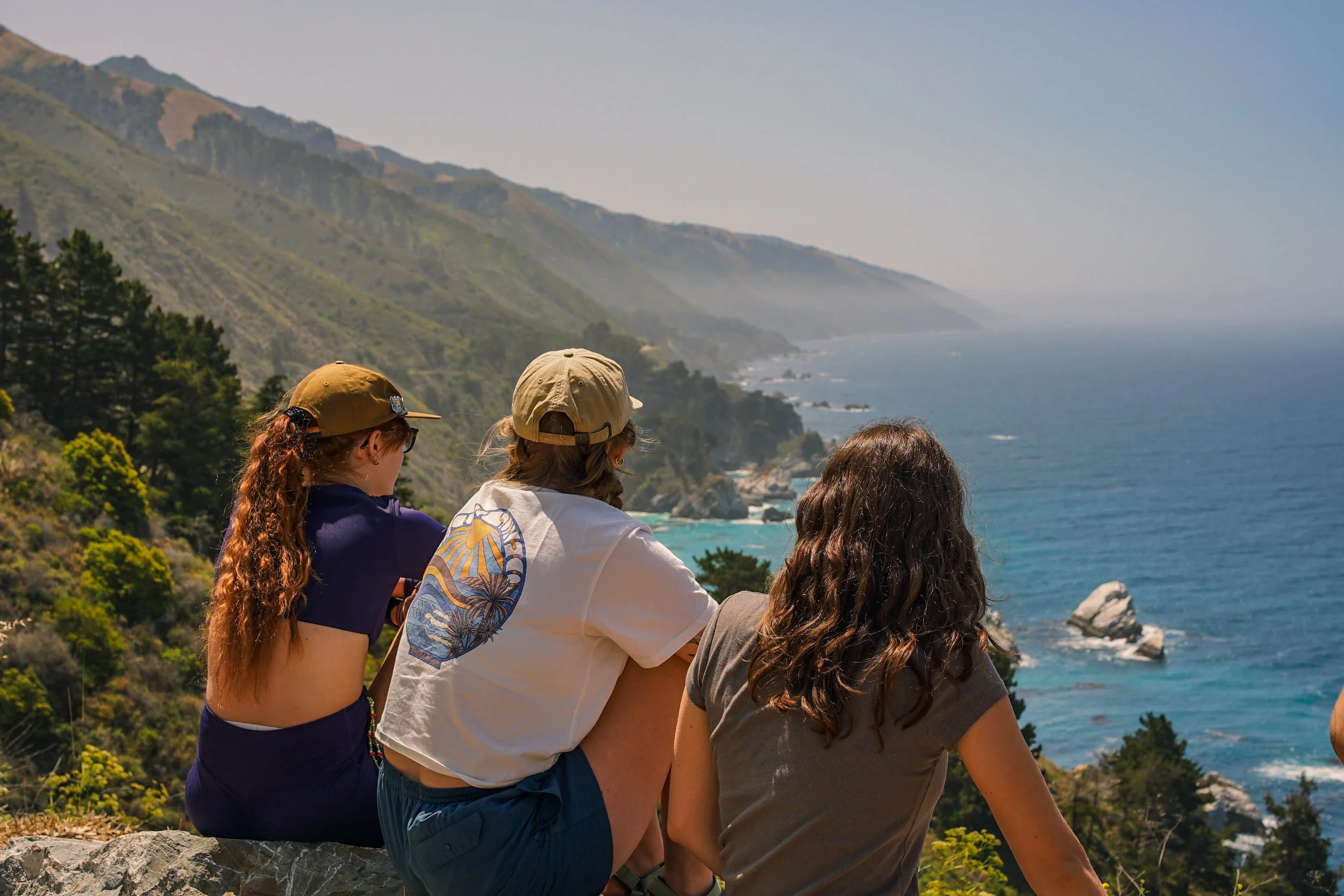 Three young women sitting on rocks overlooking a coast with cliffs, trees, and the ocean, with hills in the background under a clear sky.