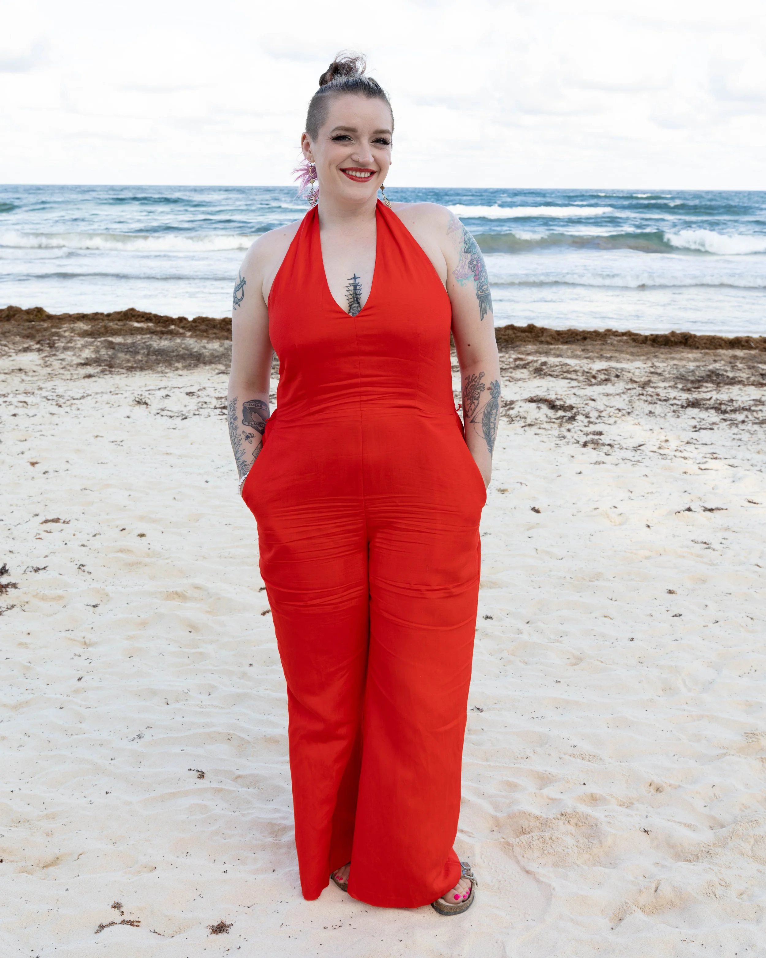 A woman in a red sleeveless dress with tattoos on her arms and chest, smiling on a sandy beach with the ocean and cloudy sky in the background.