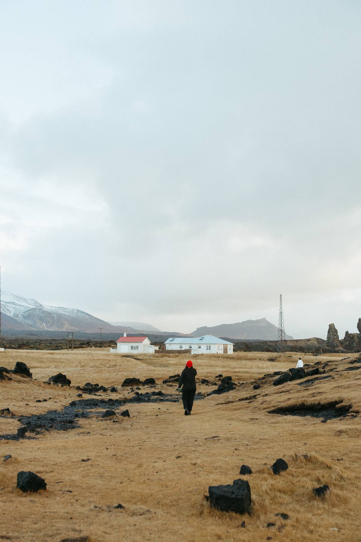 A person with a red hat walking across a yellow grassy field with scattered rocks, toward small white houses with blue roofs, mountains in the background, and a cloudy sky overhead.