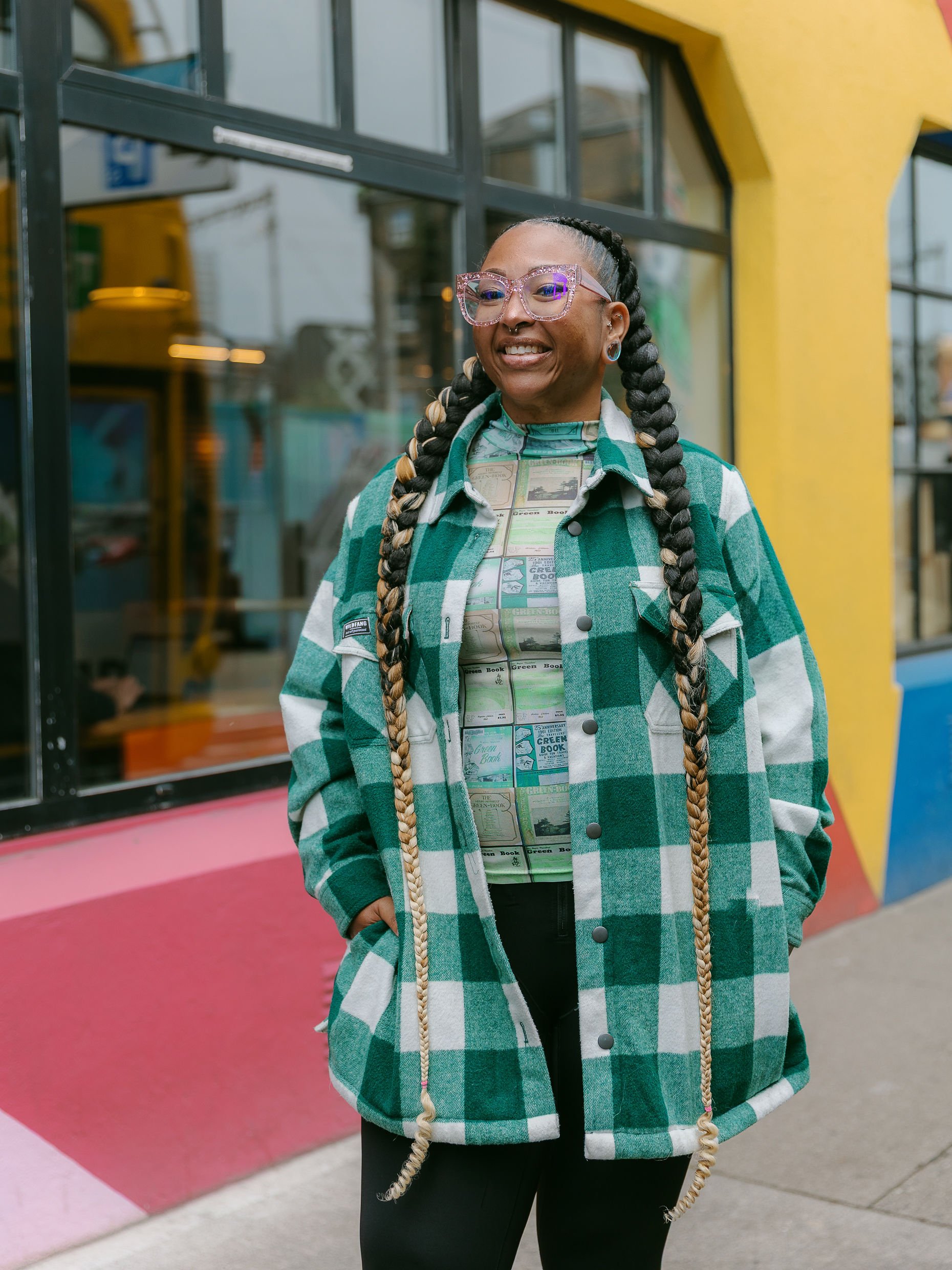 A woman with braided hair and pink glasses standing outside a colorful building.