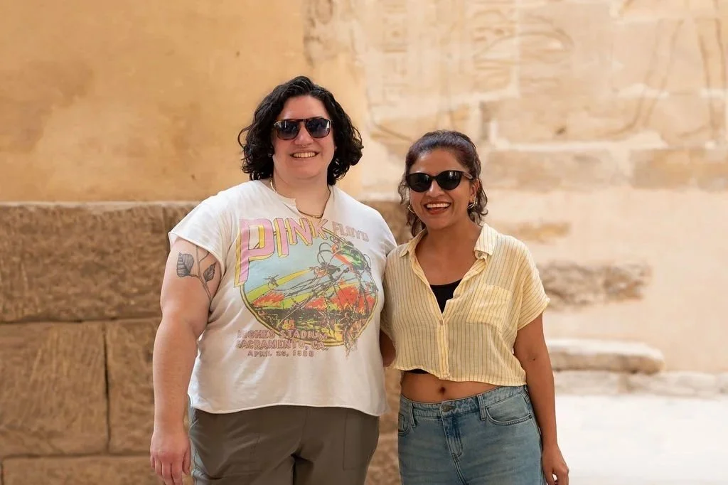 Two women with dark sunglasses smiling and standing next to each other outdoors with an ancient stone wall in the background.