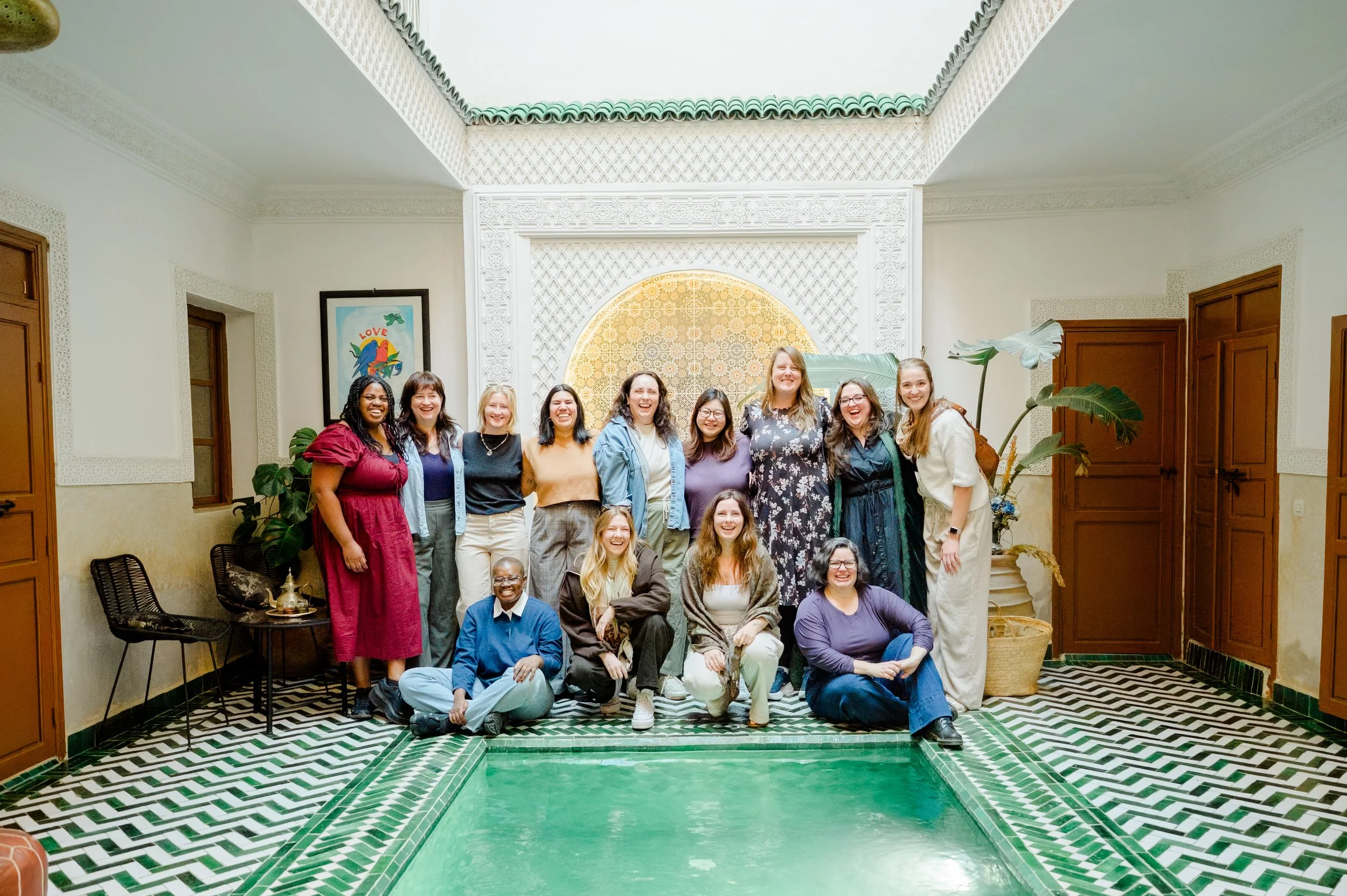 Group of diverse people posing in a decorative room with green and white floor tiles, ornate white walls, wooden accents, and tropical plants.