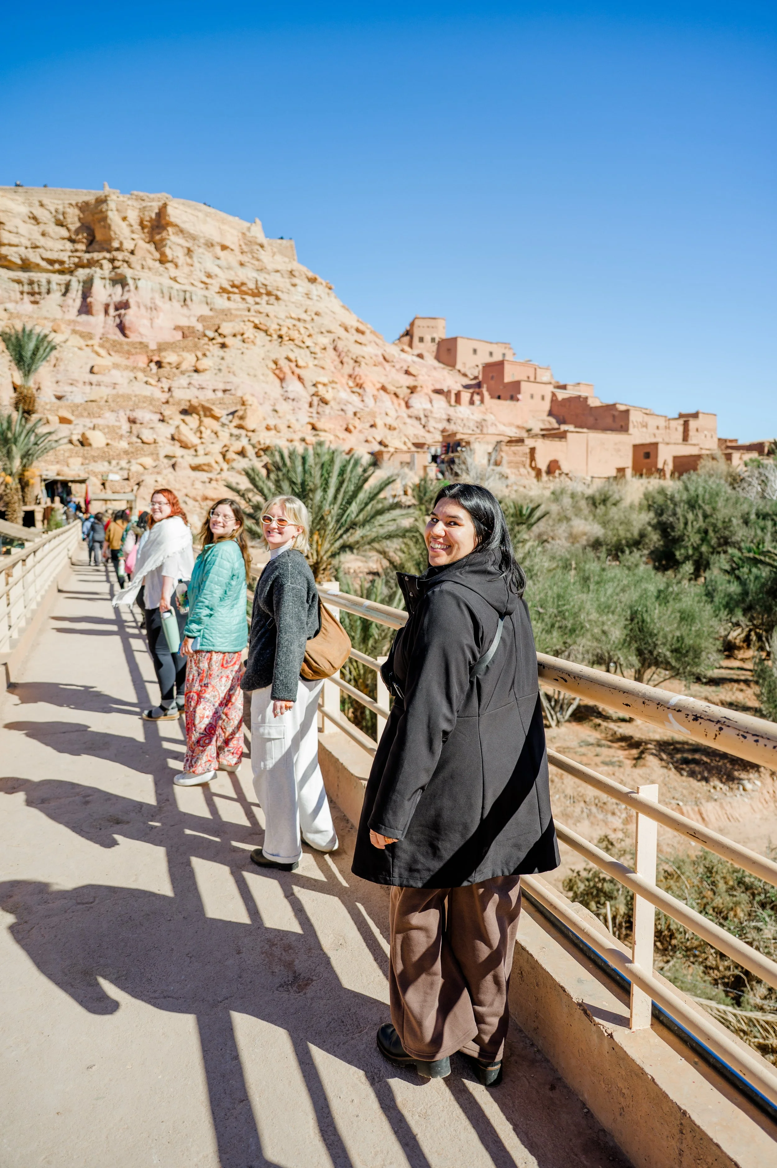 Group of smiling women standing on a walkway with a desert landscape and ancient-looking buildings on a hill in the background, under a clear blue sky.
