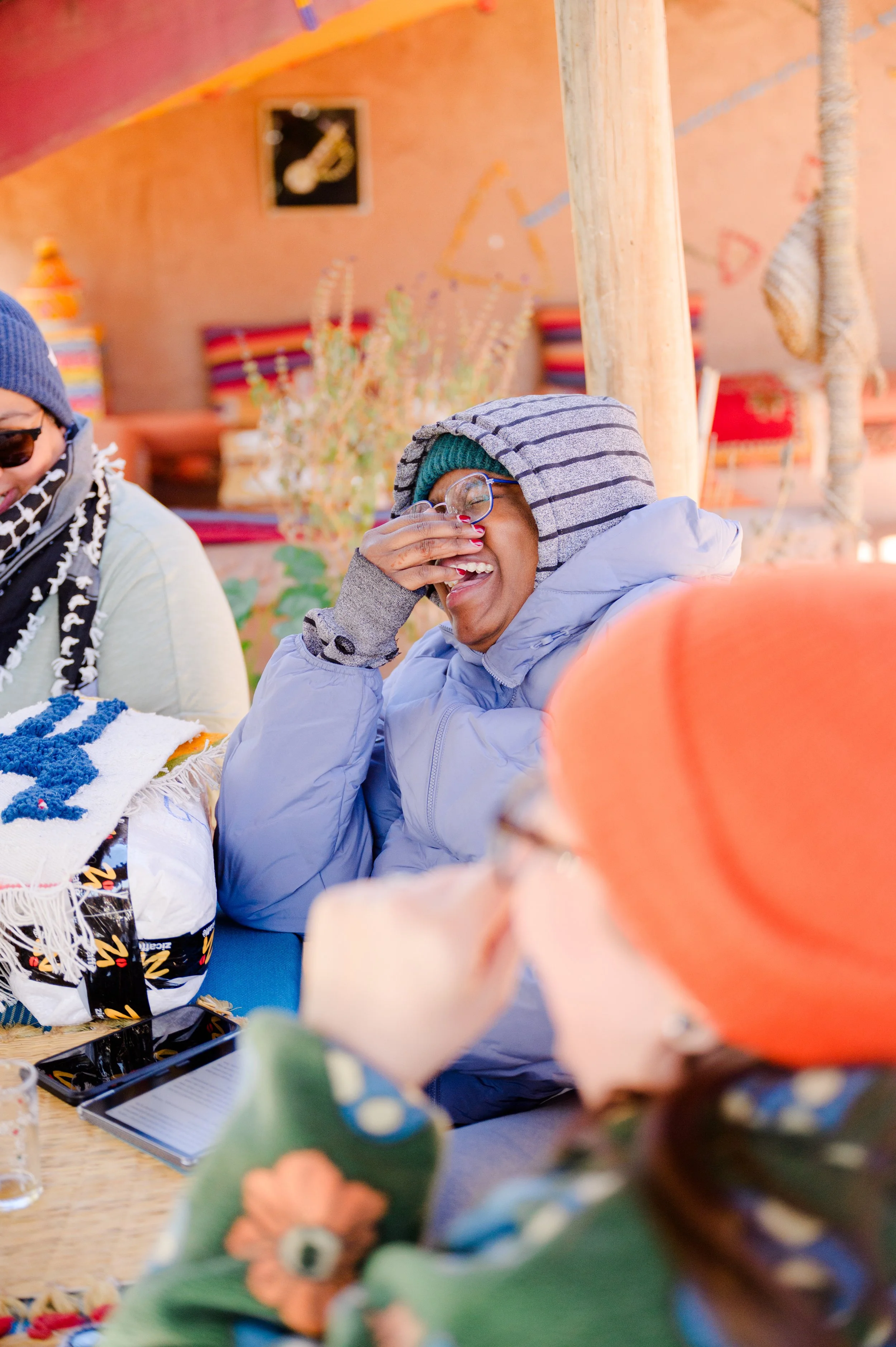 A woman wearing glasses, a green beanie, and a hoodie, laughing and covering her face with her hand, sitting at a table with others in a colorful, cozy setting decorated with textiles and artwork.