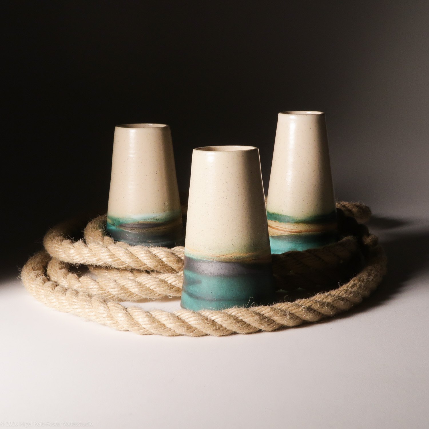 Three ceramic cups with a gradient of beige and teal green placed on a coiled beige rope, with a dark background on the left and a gradient light on the right.