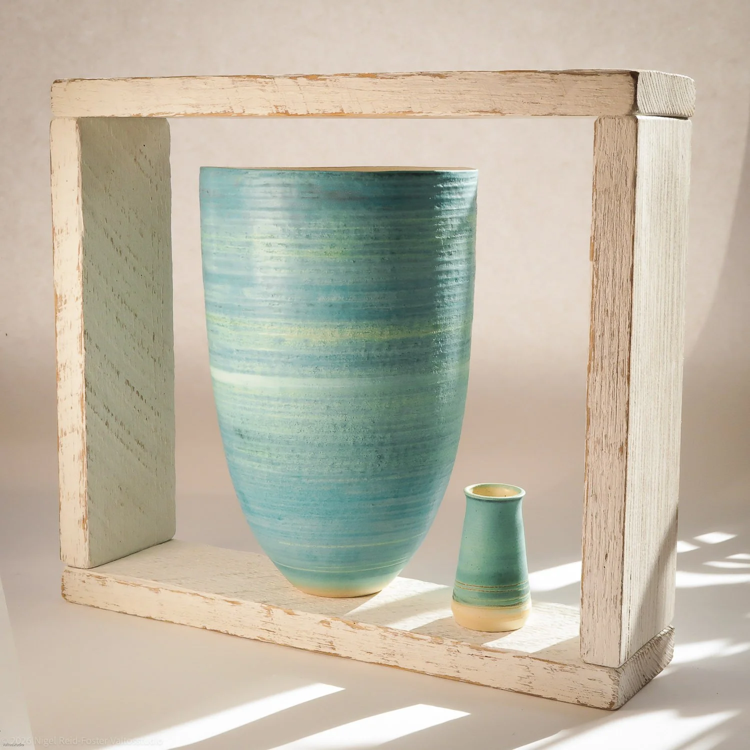 A large turquoise ceramic vase and a smaller matching vase displayed within a white wooden frame, with soft lighting creating shadows.