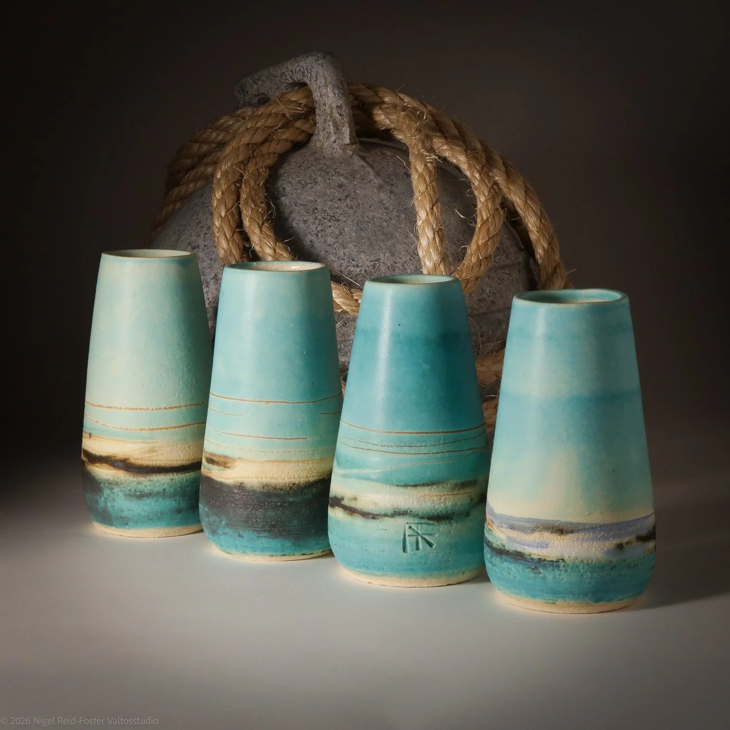 Four ceramic vases with ocean or beach landscape designs, arranged in a row in front of a large stone with a rope attached to it, against a dark background.