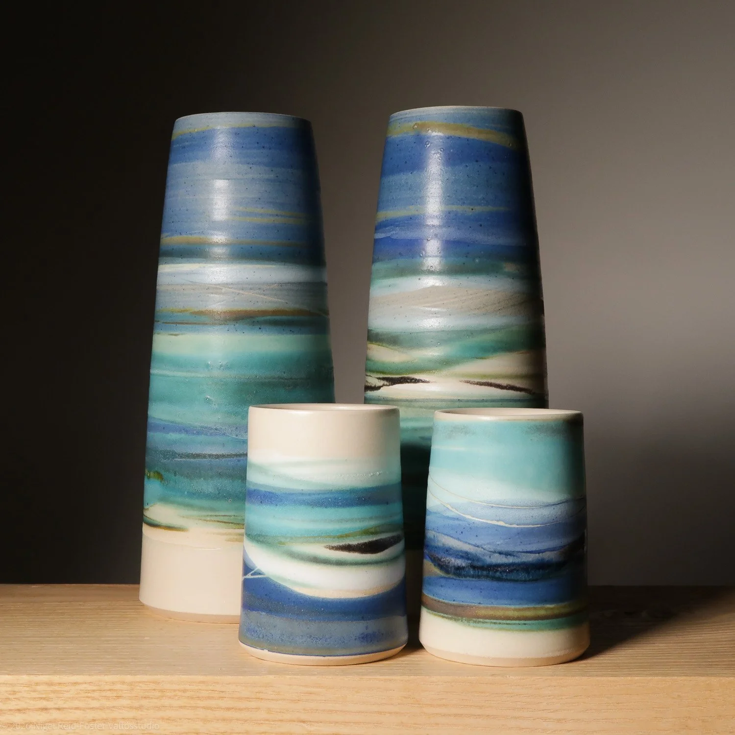 Four ceramic vases with blue, green, beige, and black horizontal streaks, arranged on a wooden surface against a dark background.