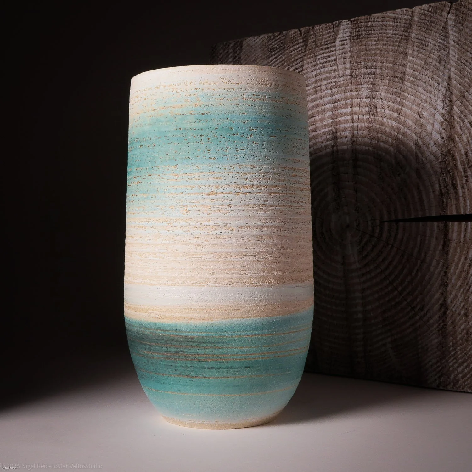 A cylindrical ceramic vase with horizontal layers of white, beige, light blue, and turquoise colors, placed on a surface against a dark background with a wooden log behind it.