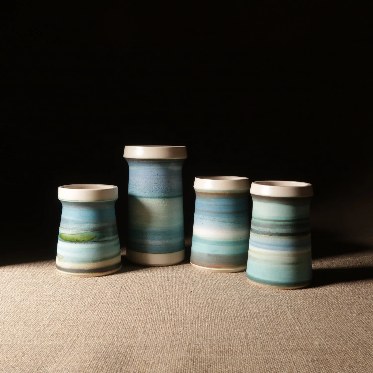 Four ceramic cups with blue and white striped glaze, arranged on a beige surface with a dark background.
