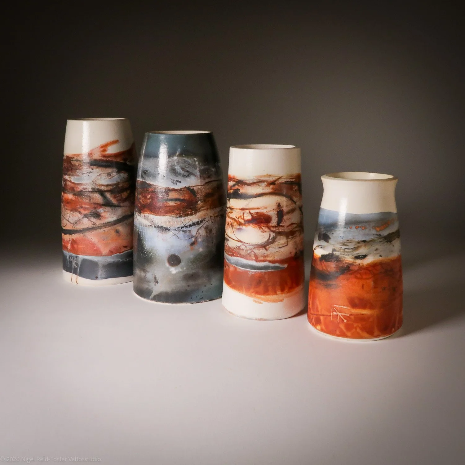 Four ceramic vases with abstract marbled patterns in shades of orange, black, gray, white, and cream, arranged in a line on a white surface with a dark background.