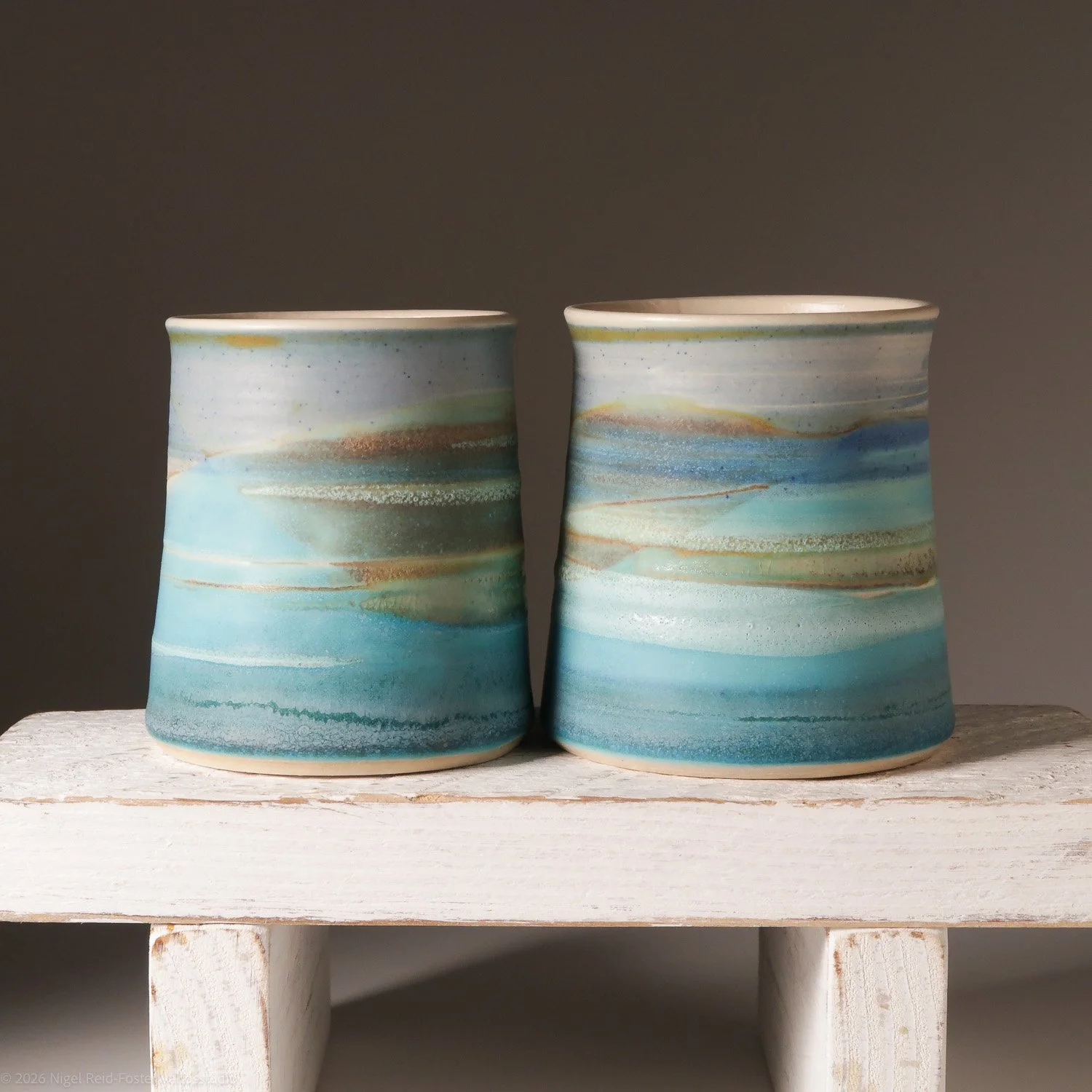 Two ceramic vases with blue, green, beige, and brown watercolor-like patterns sitting on a rustic white wooden surface against a simple dark background.