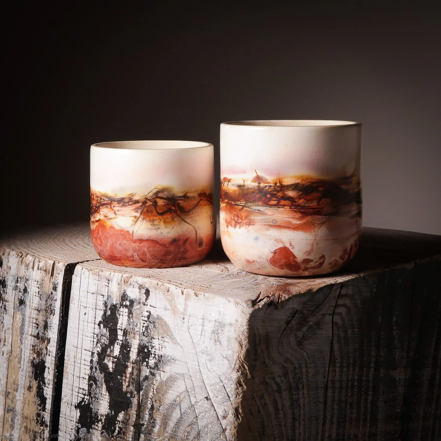 Two ceramic cups with abstract marbled designs in shades of red, brown, and black, resting on a weathered wooden surface.