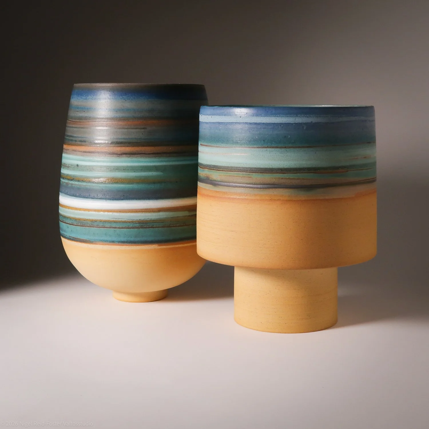 Two ceramic vases with horizontal striped patterns in shades of blue, teal, cream, and orange, placed on a white surface with a dark background.
