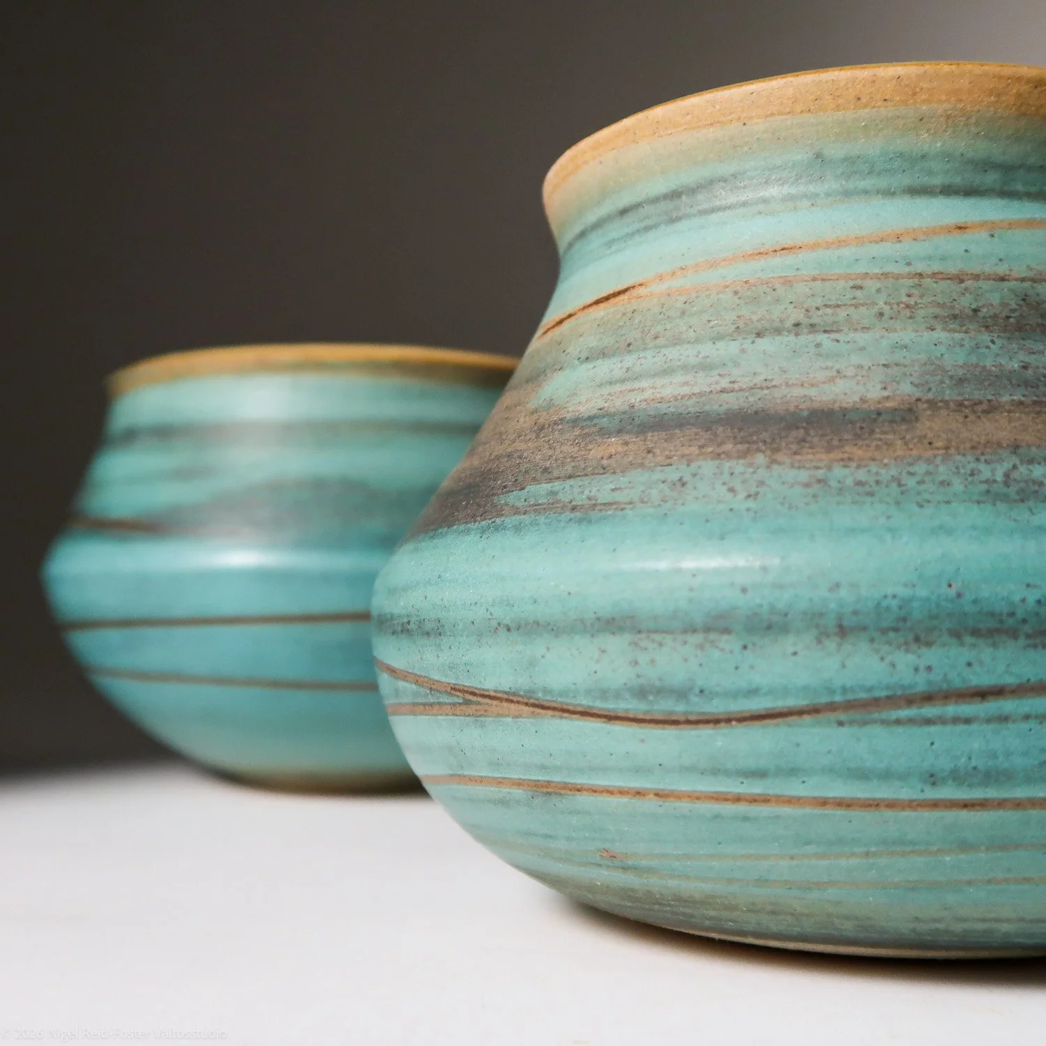 Two ceramic vases with turquoise and brown horizontal stripes, set against a dark background.