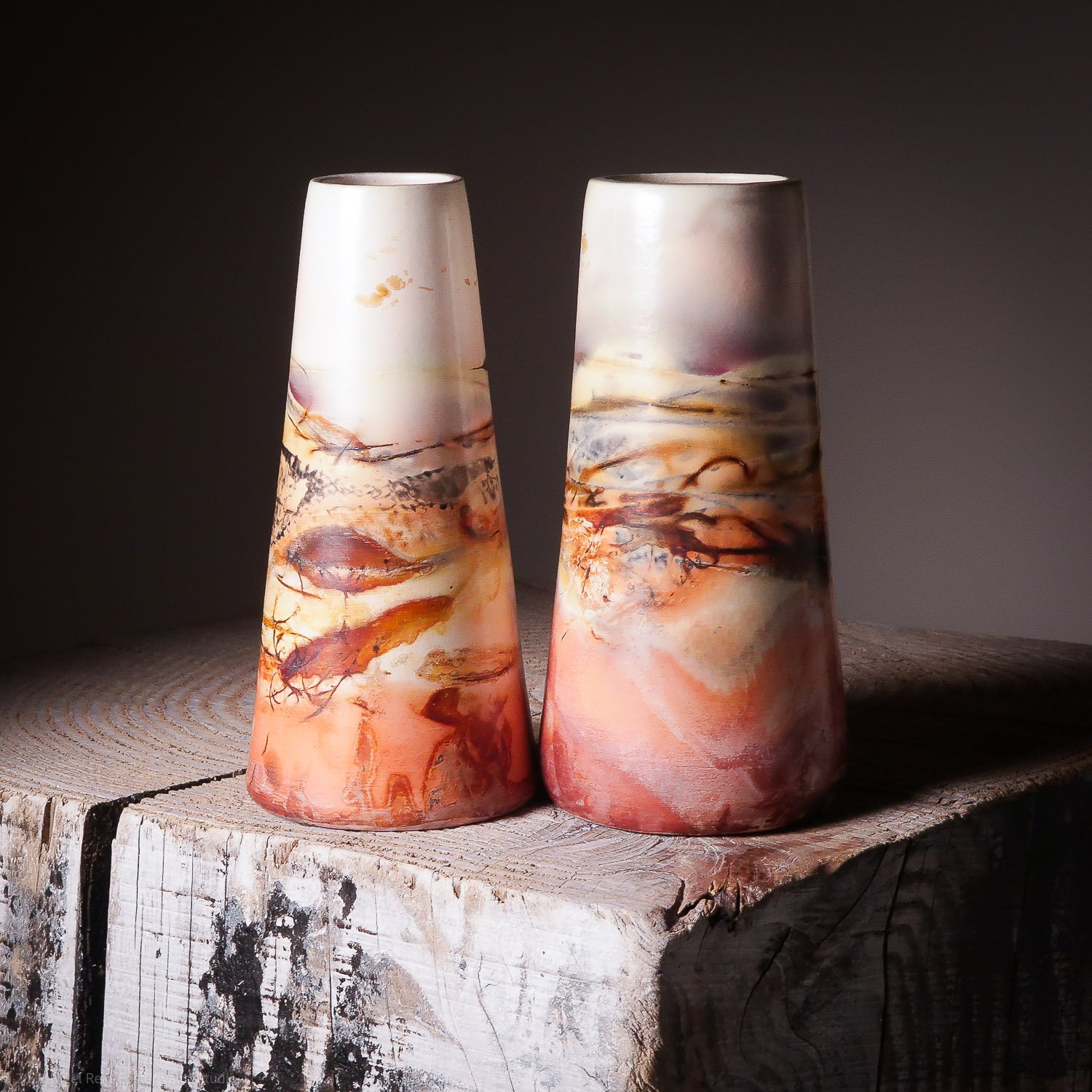 Two tall, conical glass vases with abstract orange, pink, and dark swirl patterns, placed on a weathered wooden surface with a dark background.