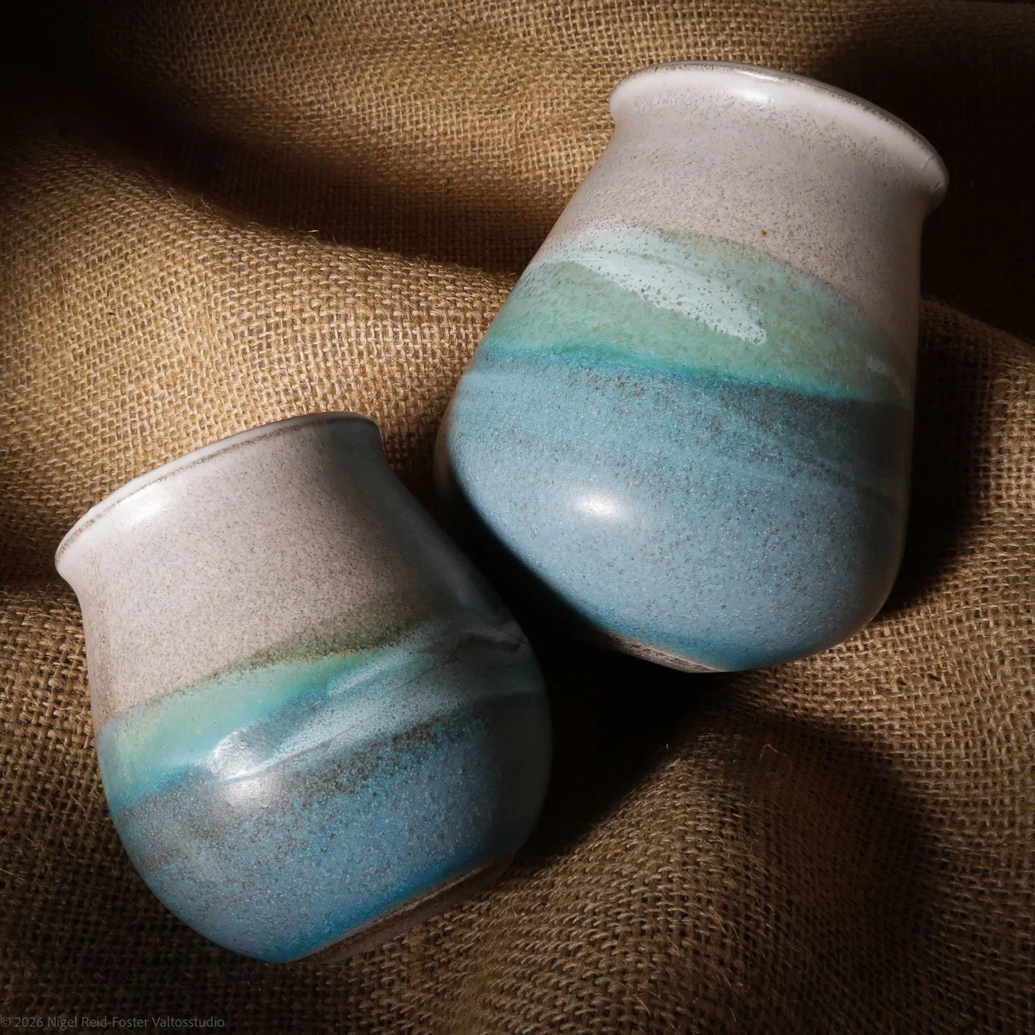 Two ceramic vases with a layered, multicolored glaze in shades of blue, green, and tan, resting on a textured brown cloth.