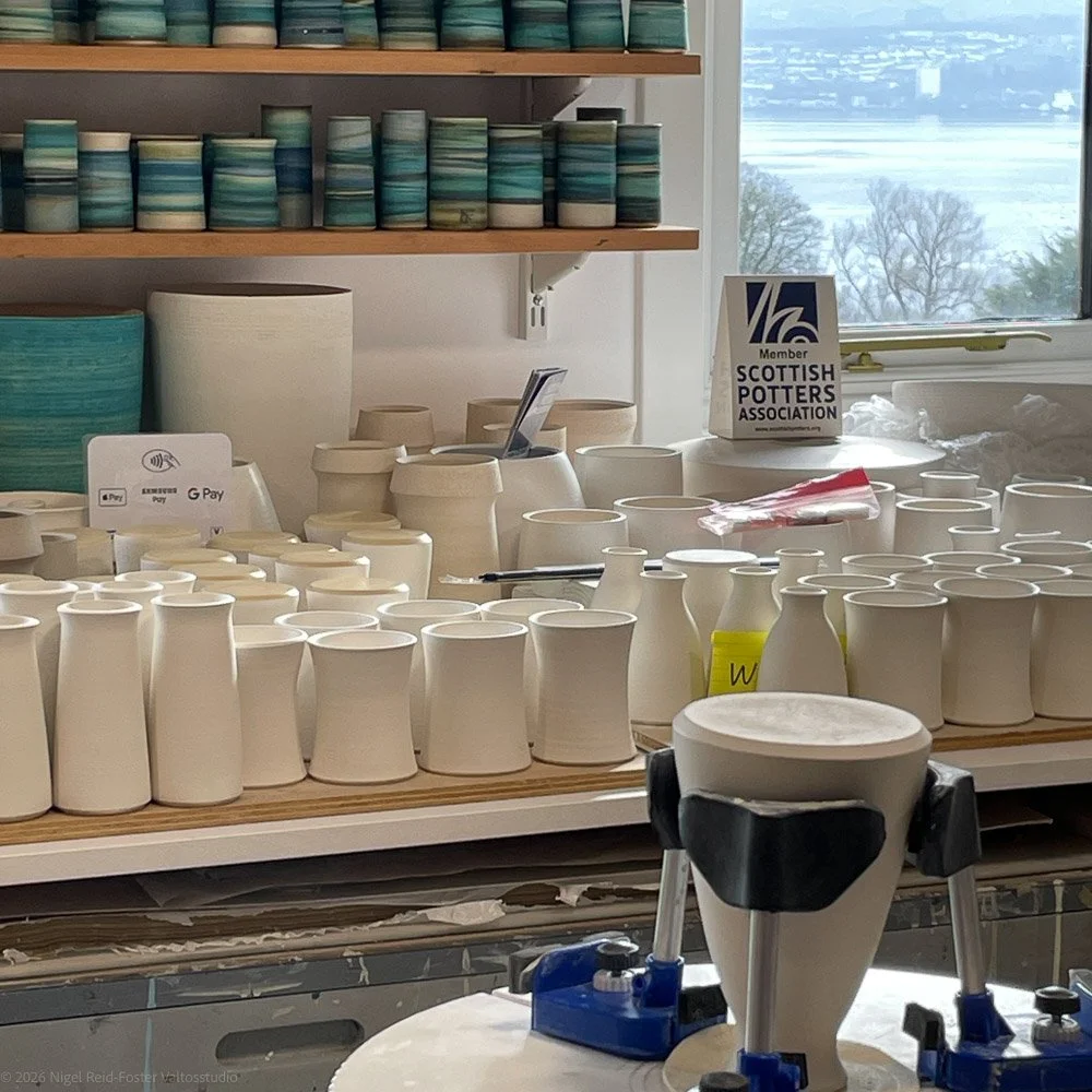 A pottery studio shelves filled with various glazed ceramic cups and vessels, with unglazed pottery in the foreground and a window overlooking a body of water and trees outside.