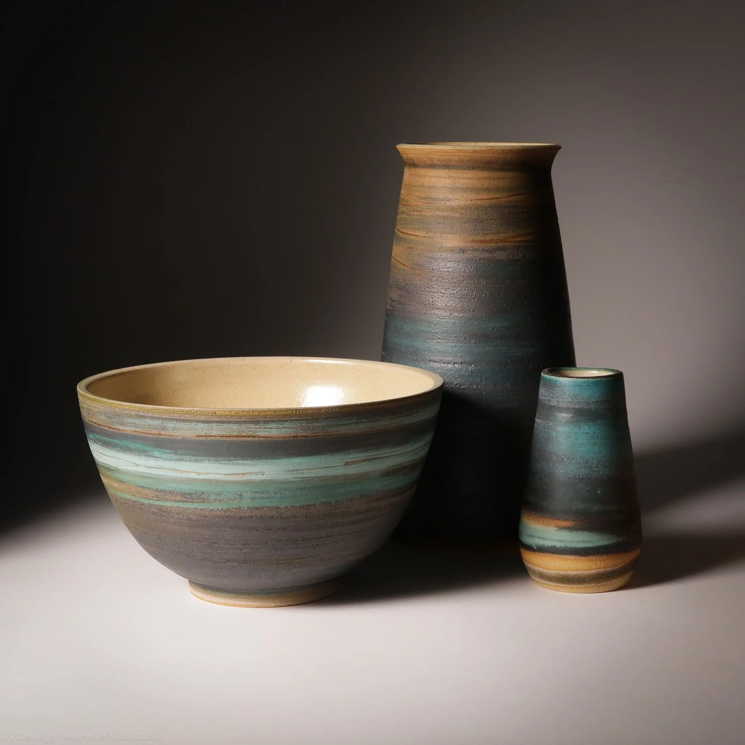 Three ceramic vessels: a large bowl, a tall tapered vase, and a smaller rounded vase, all with earthy-colored, striped glaze, arranged on a white surface against a dark background.
