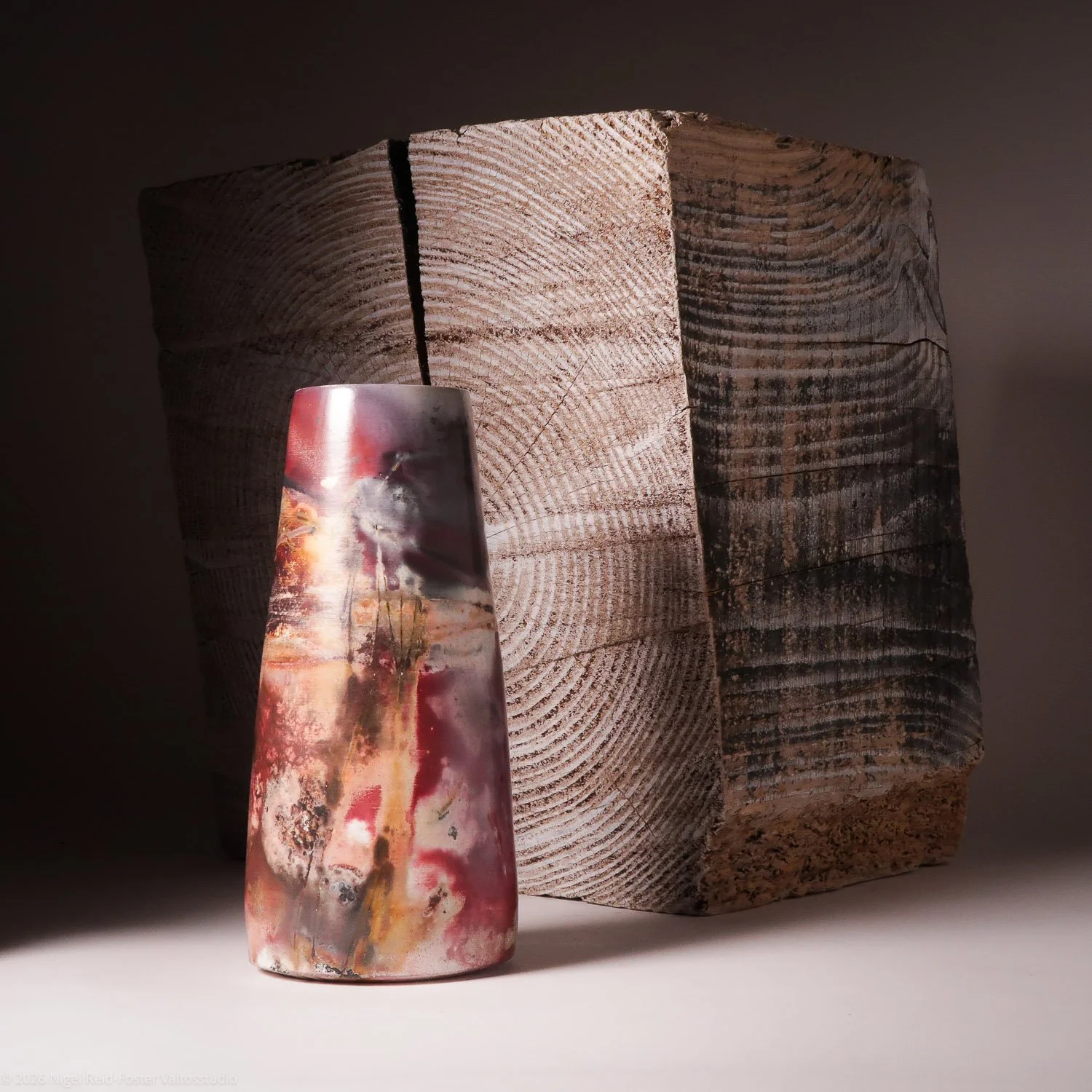 A tall, abstract, multicolored ceramic vase with pink, red, cream, and dark streaks, placed in front of a large, rough, wooden block with visible grain and circular growth rings.