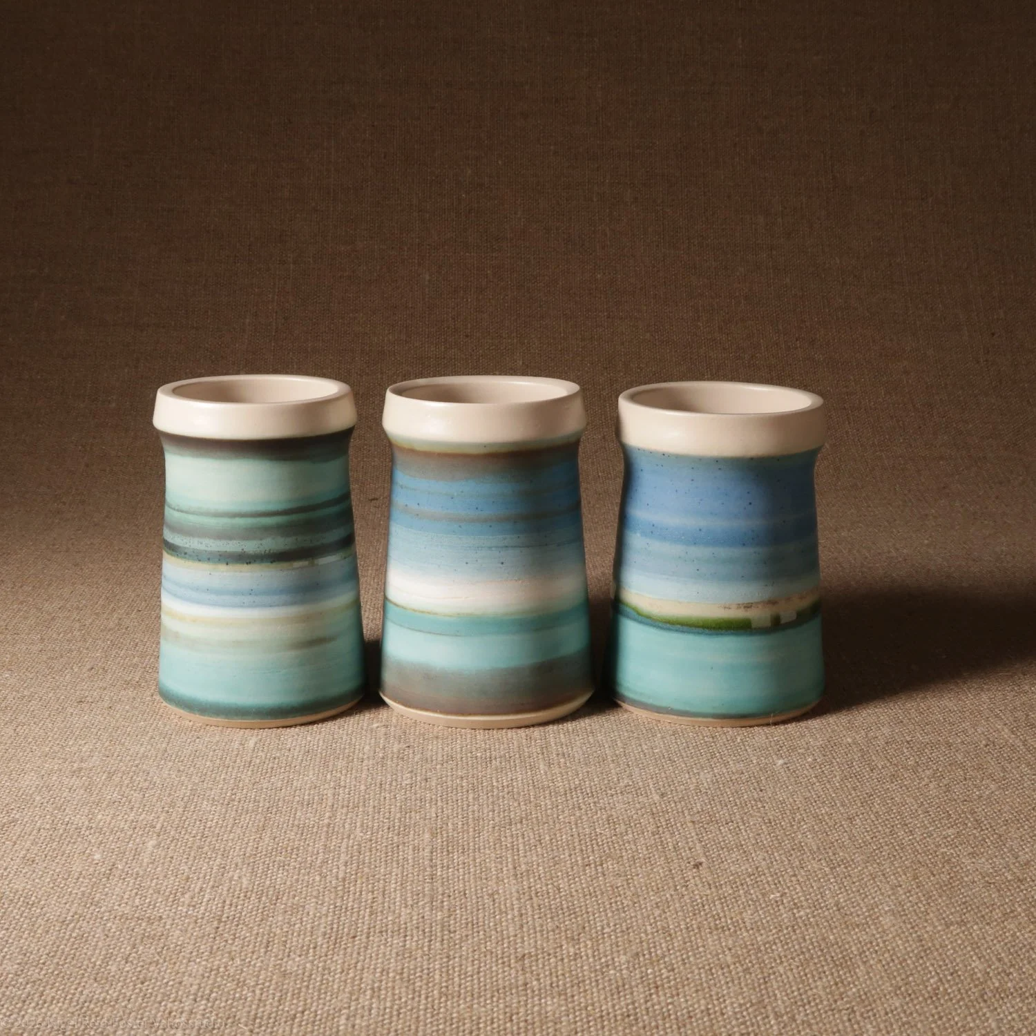 Three ceramic vases with blue and green horizontal stripe patterns on a beige surface, against a brown background.