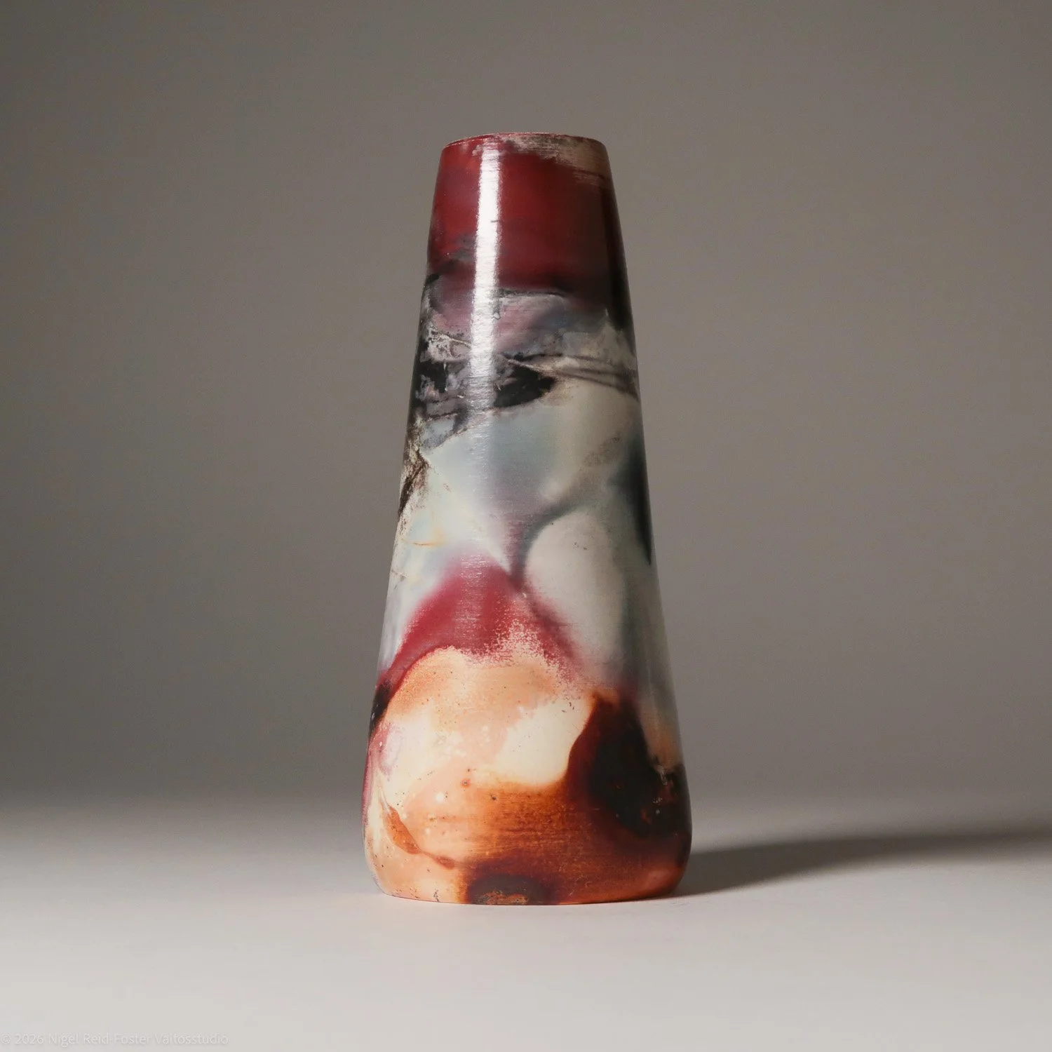 A tall, tapered glass vase with abstract multicolored patterns in shades of red, white, black, and beige, placed on a plain surface against a plain, neutral gray background.