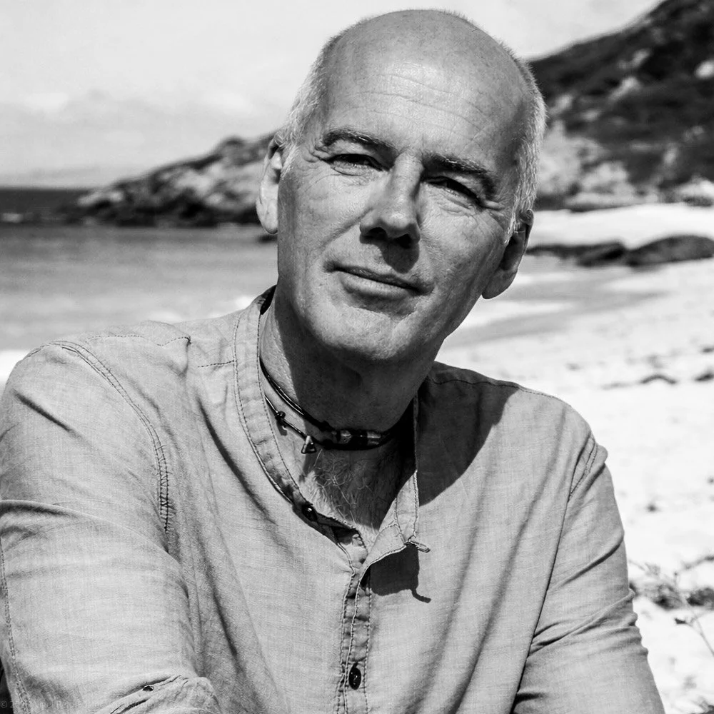 A black-and-white photo of a man with a shaved head smiling outdoors, with mountains and snow in the background.
