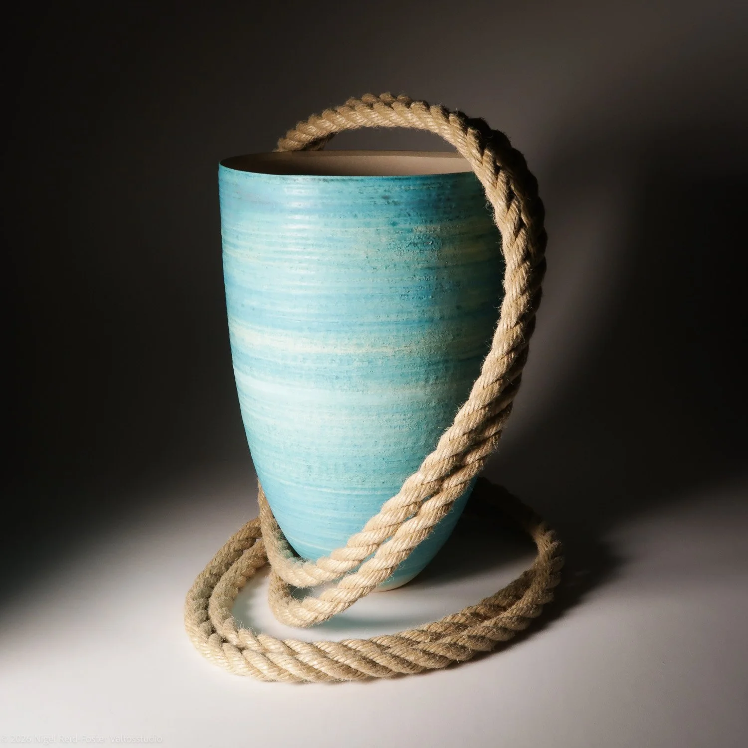 A large blue ceramic vase with a natural fiber rope handle draped around it, placed on a surface with a dark background.