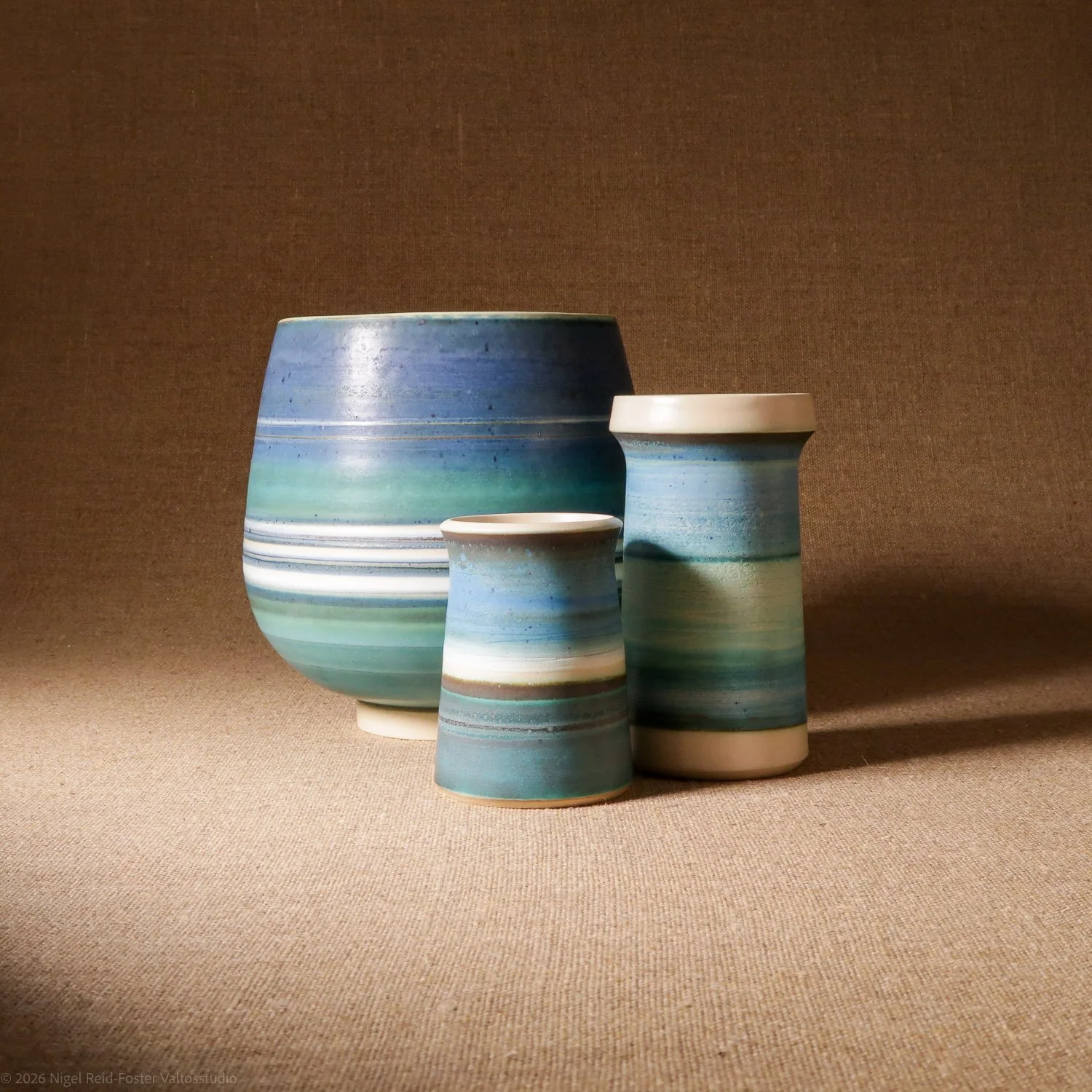 Three ceramic vases with a blue, green, and white striped glaze on a neutral textured surface against a brown background.
