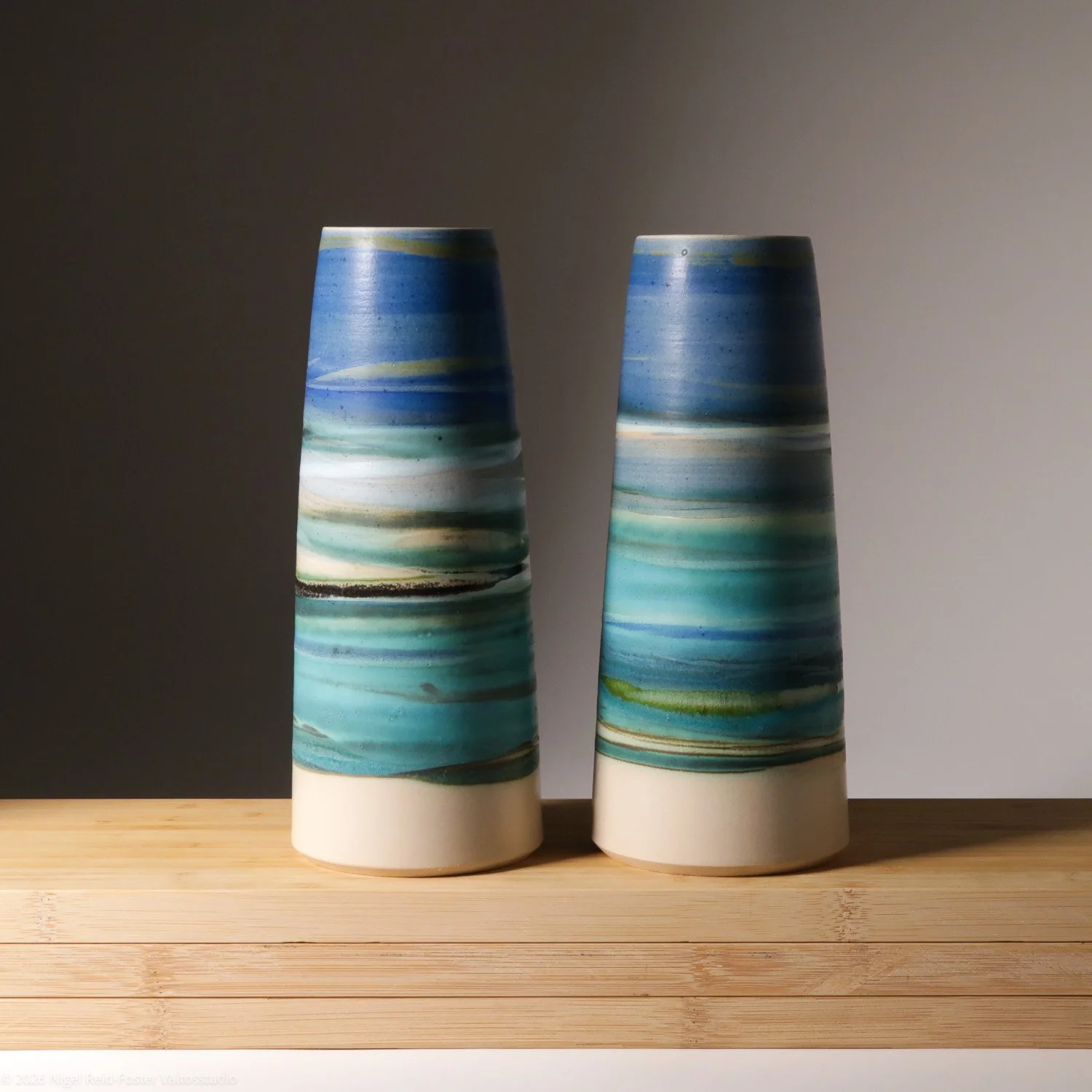 Two tall ceramic vases with blue, green, and white horizontal stripes, placed on a wooden surface against a neutral background.