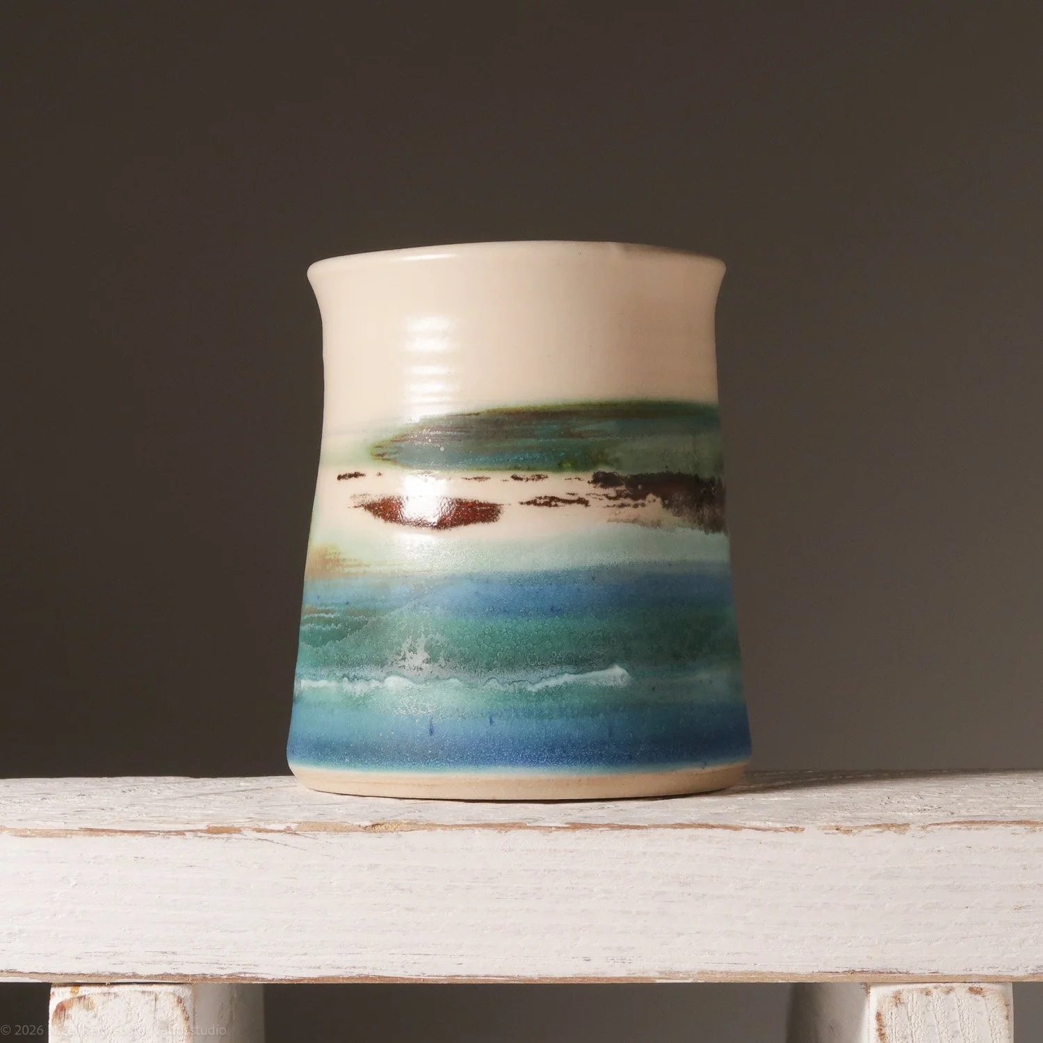 Ceramic vase with a landscape painting featuring water, land, and sky, placed on a white wooden surface against a dark background.