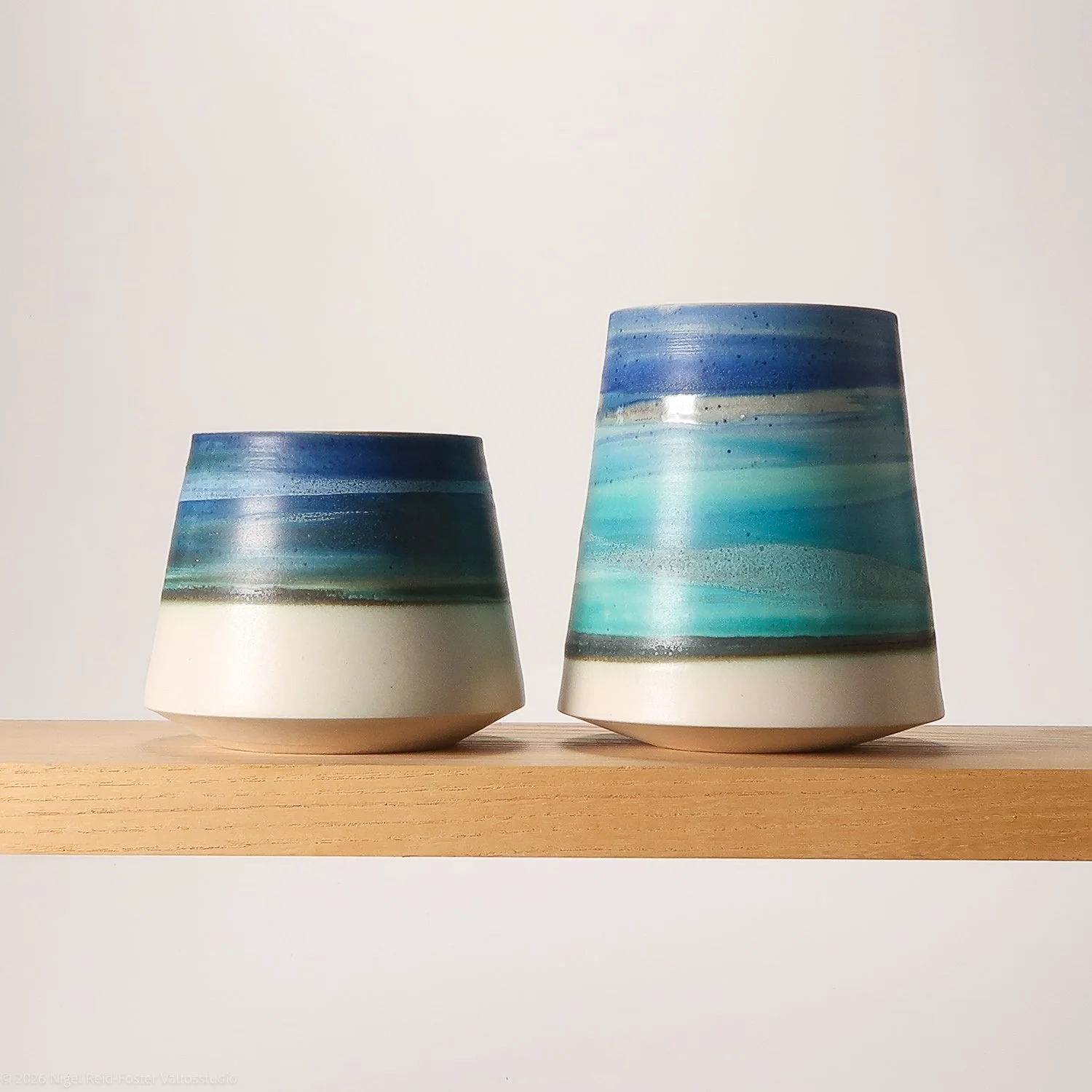 Two ceramic cups with blue and teal gradient glaze patterns, placed on a wooden shelf against a plain white background.