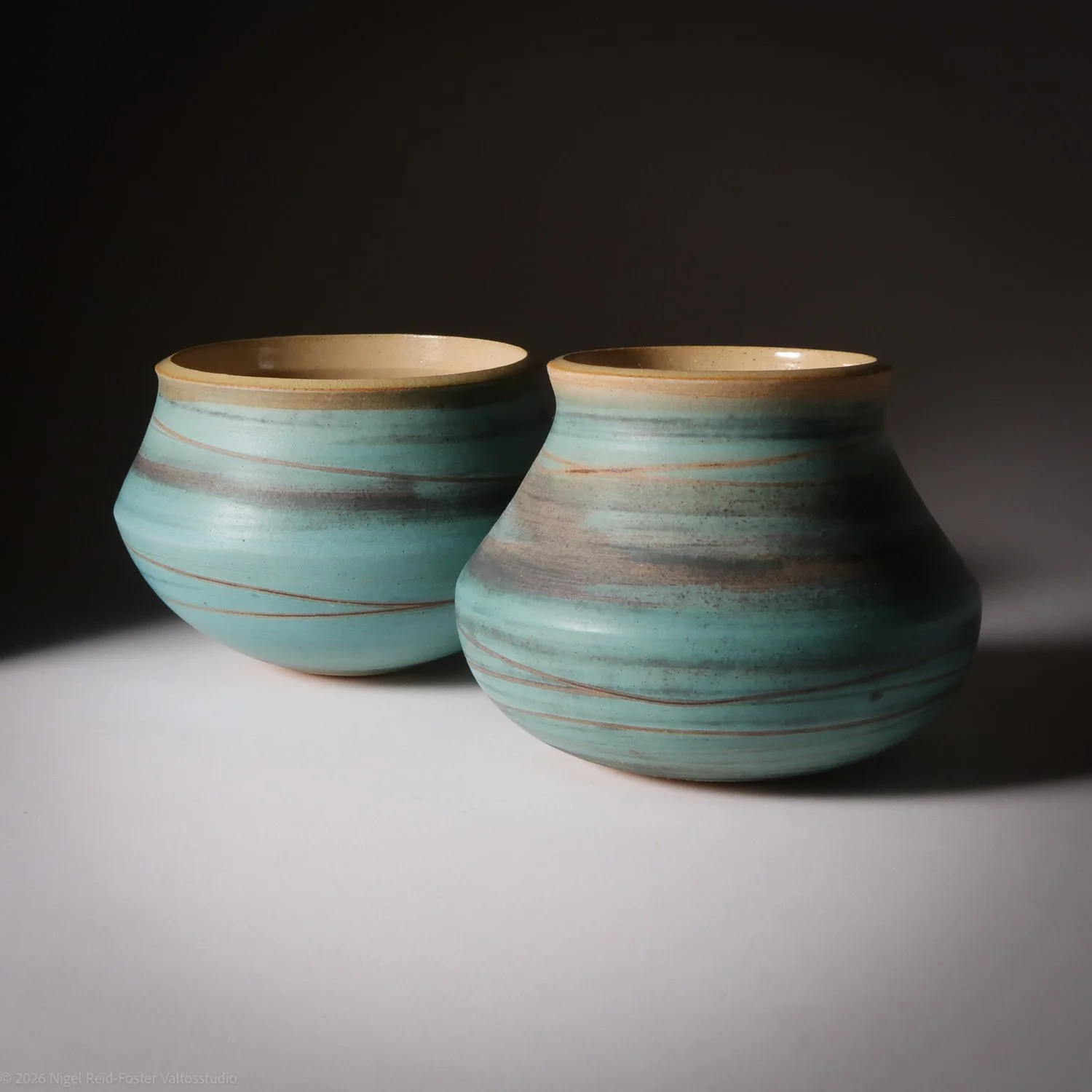 Two ceramic vases with a rounded, bulbous shape and horizontal striped patterns in shades of blue and brown, placed on a white surface with a dark background.
