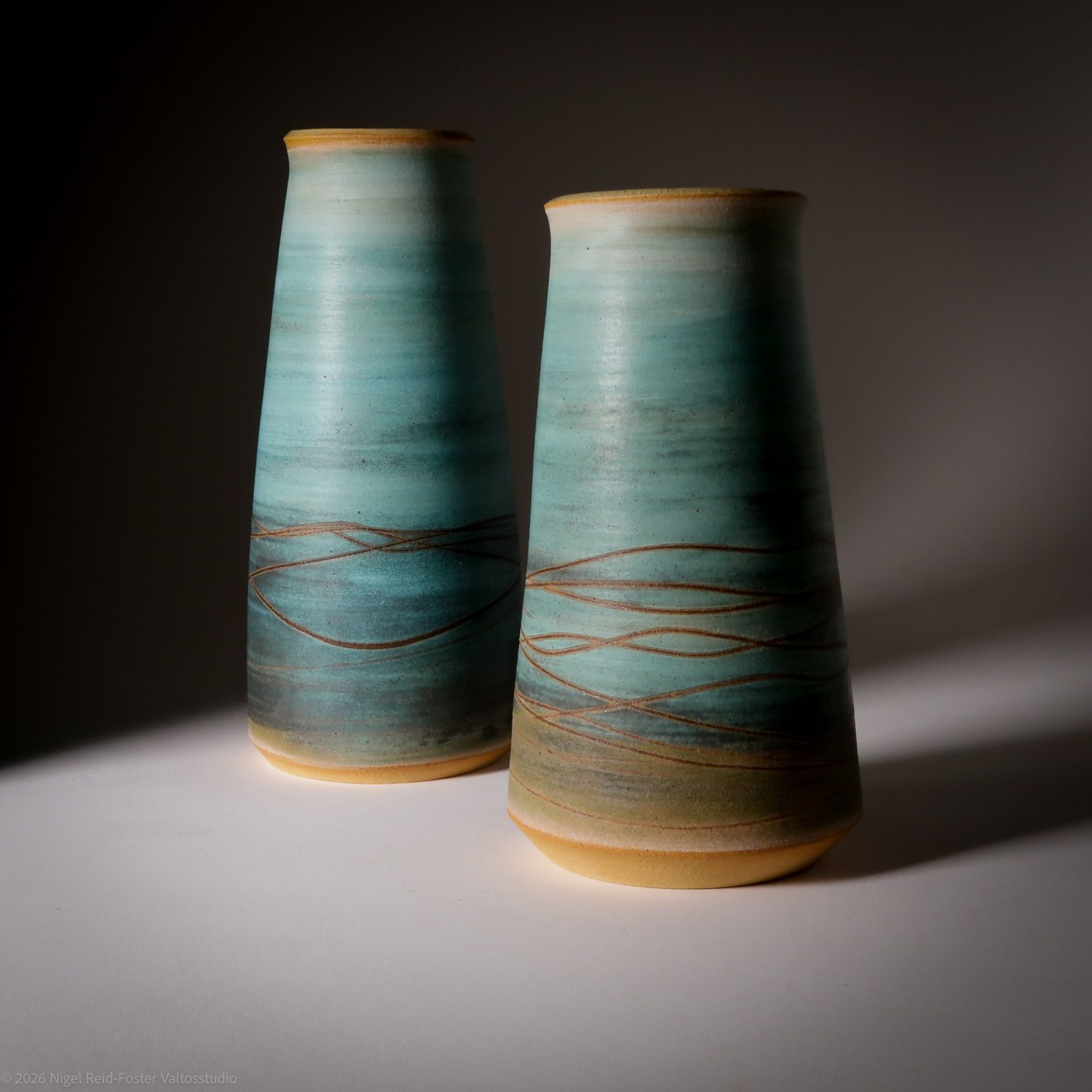 Two ceramic vases with a blue-green glaze and brown line designs, placed on a gray surface with shadows.