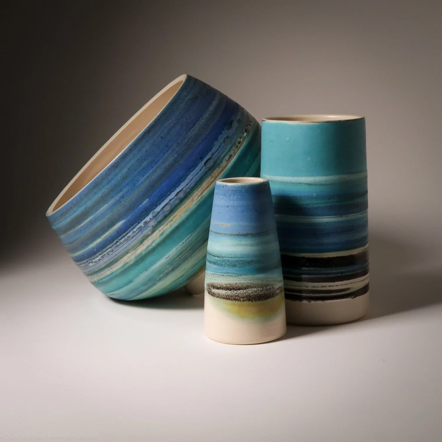Three ceramic vases with blue and green watercolor-like horizontal stripe patterns, one lying on its side and two standing upright, against a neutral background.