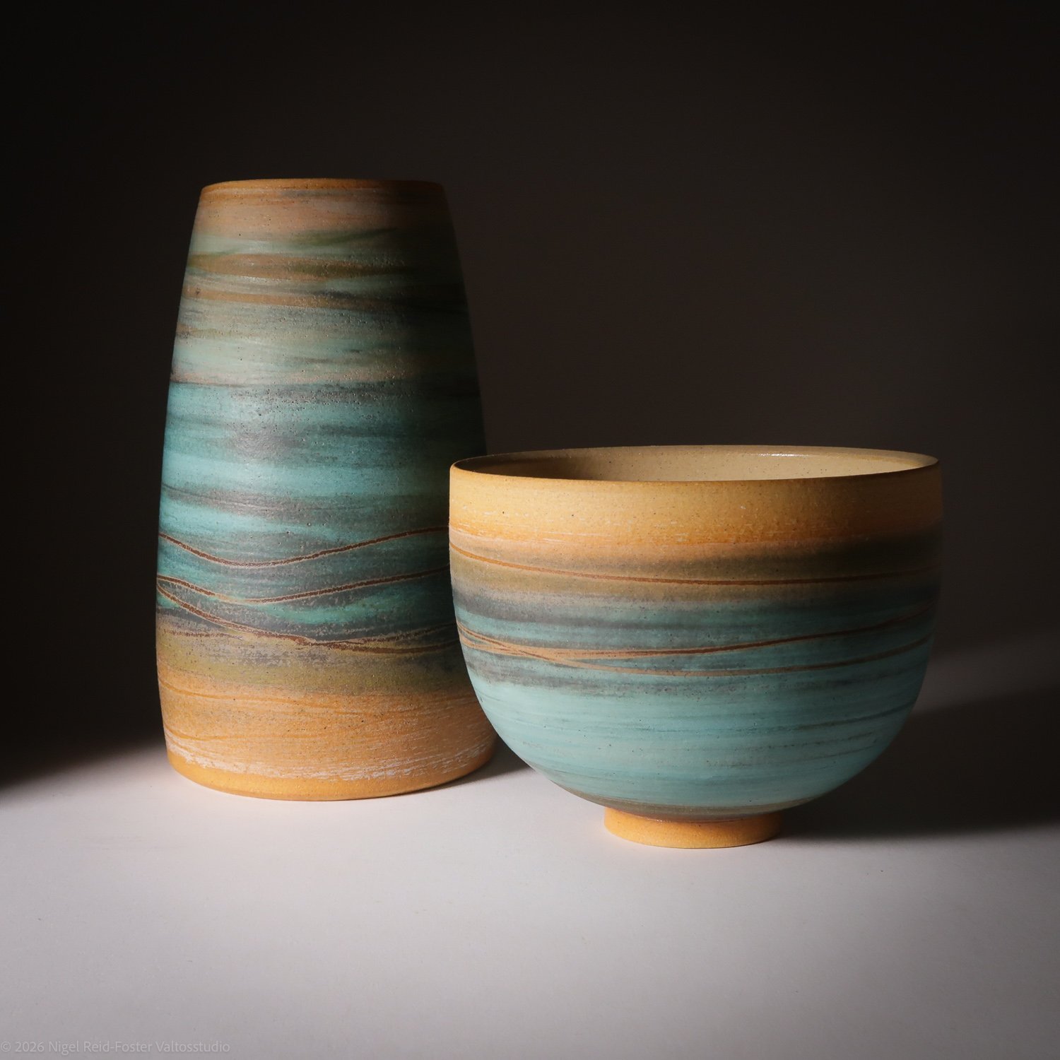Two ceramic vases with earth-tone and turquoise striped glaze lighting, one taller and cylindrical, the other shorter and round, against a dark background.