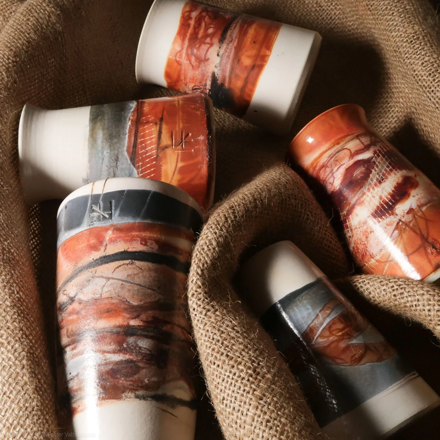Group of ceramic mugs with abstract landscape paintings, placed on a burlap cloth.