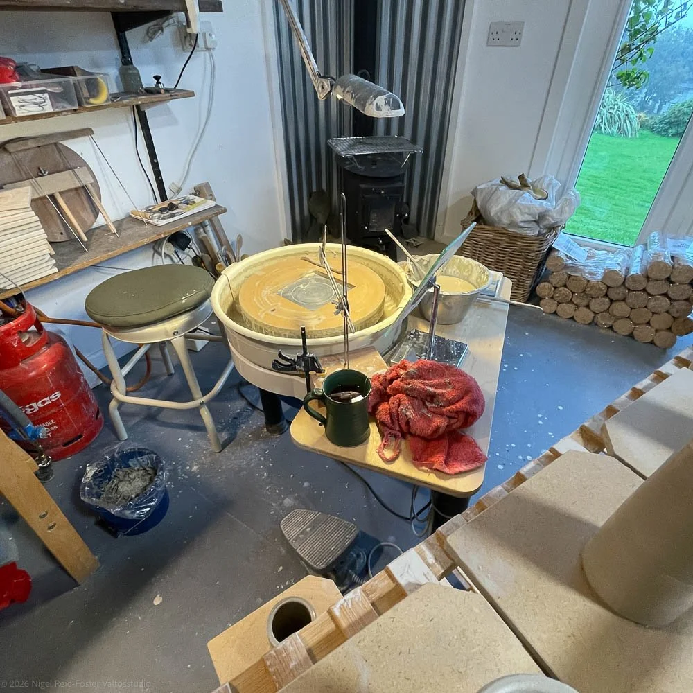 A pottery wheel with clay spinning on it in a pottery studio. Various pottery tools, a mug, and a red cloth are on the table nearby. Shelves with supplies and a wood stove are visible in the background, along with a window showing a green outdoor area.