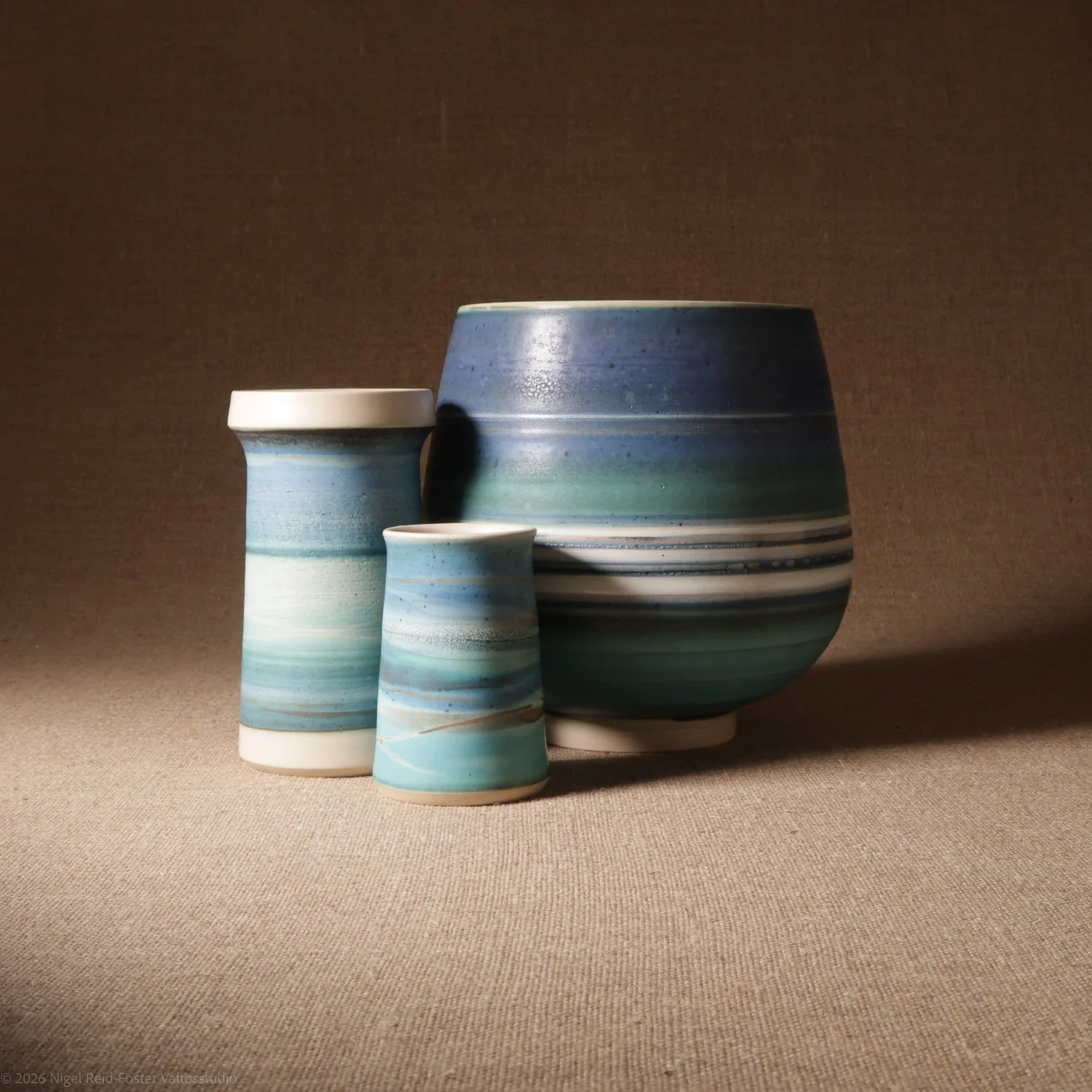 Three striped ceramic vases of varying sizes in shades of blue, teal, and white, displayed against a neutral background.