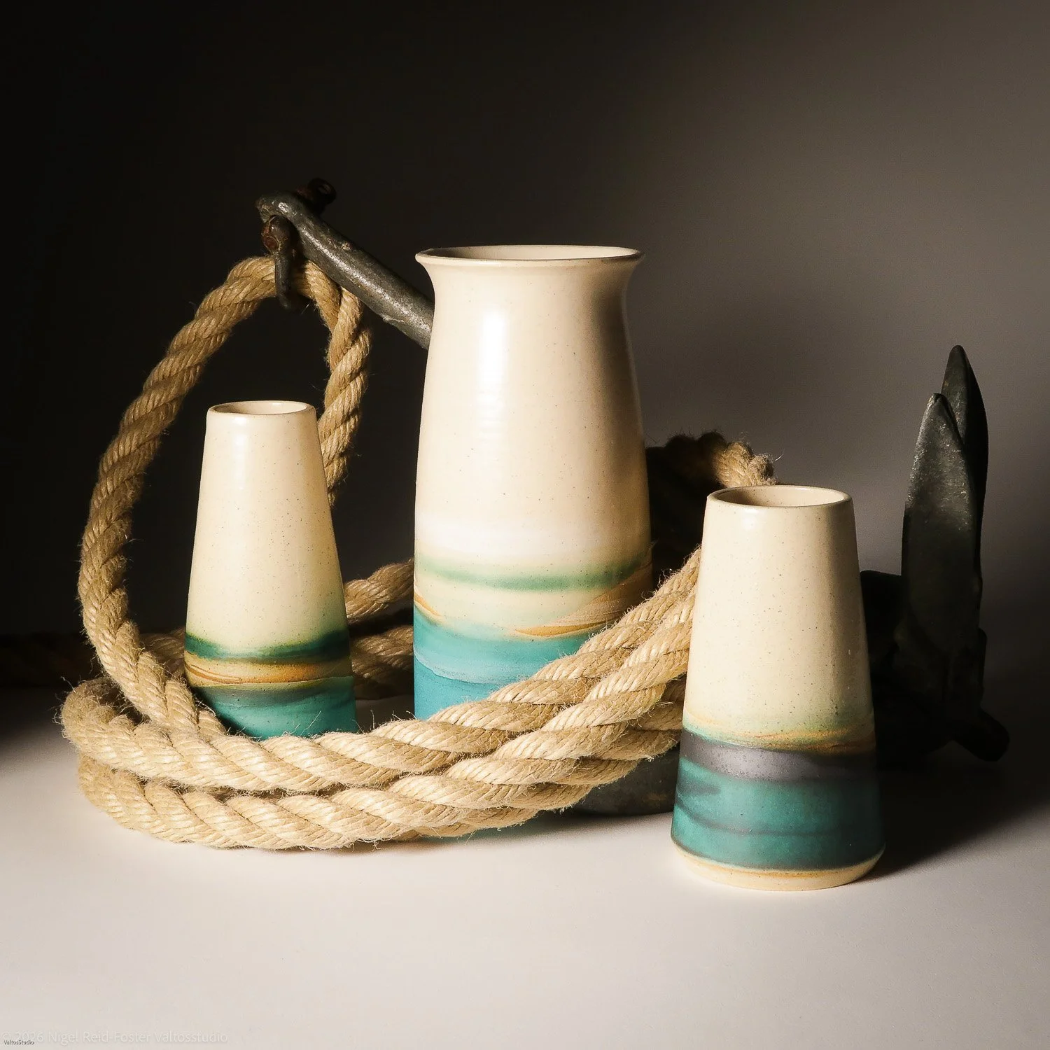 Ceramic vases with beach-like glaze in shades of green, blue, and beige, arranged with a thick rope and metal tools on a dark background.