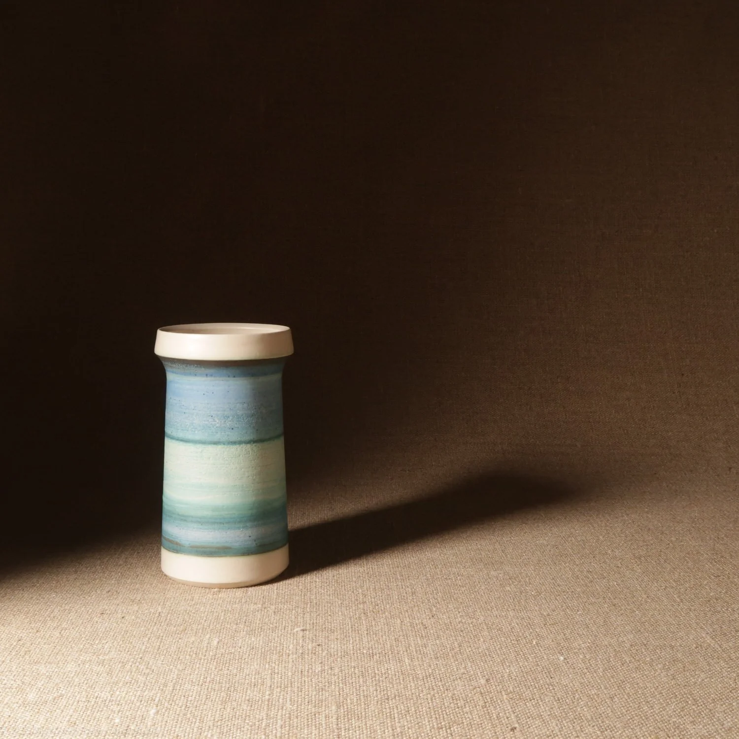 A ceramic vase with blue and white horizontal stripes, placed on a light-colored surface with a dark brown background, casting a shadow.
