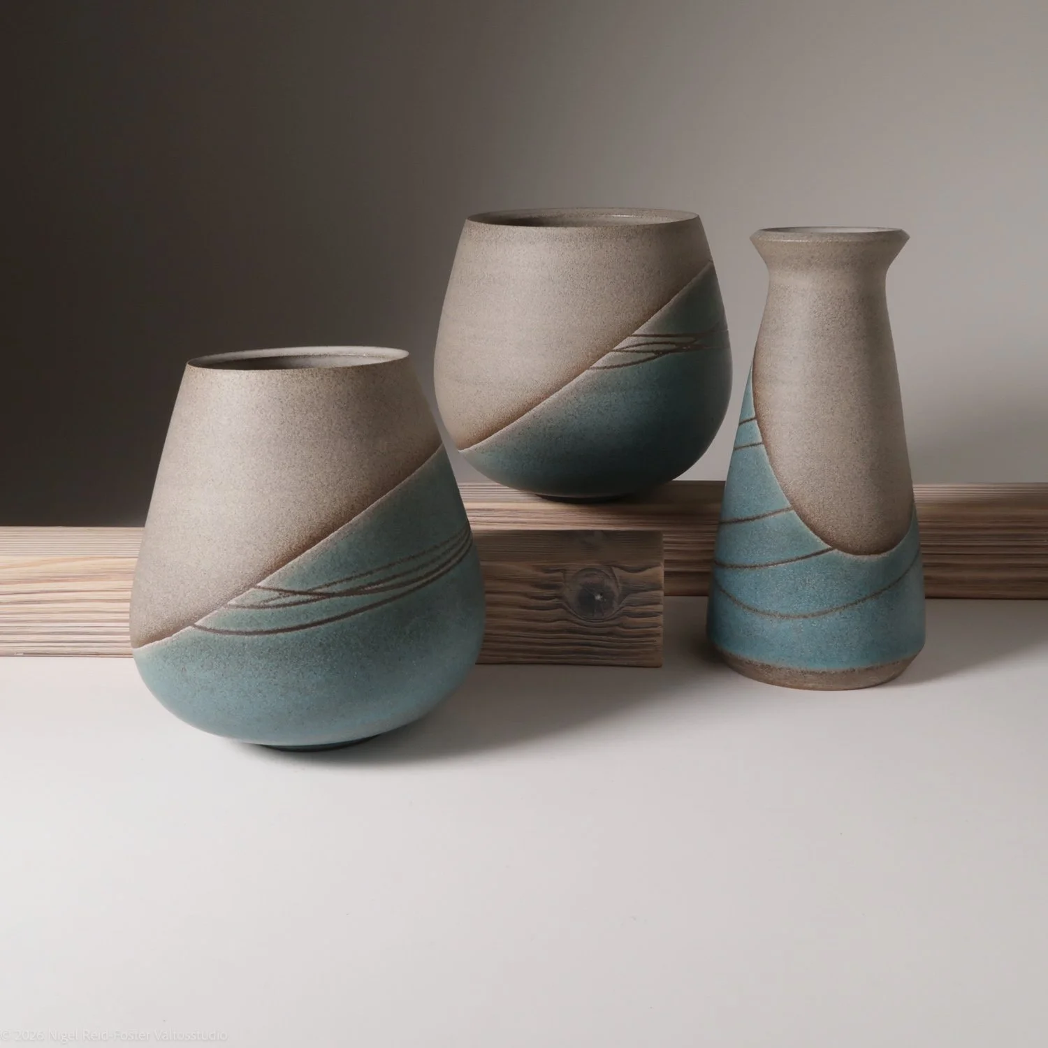Three ceramic vases with beige and blue color patterns, arranged on a wooden plank and white surface against a gradient gray background.