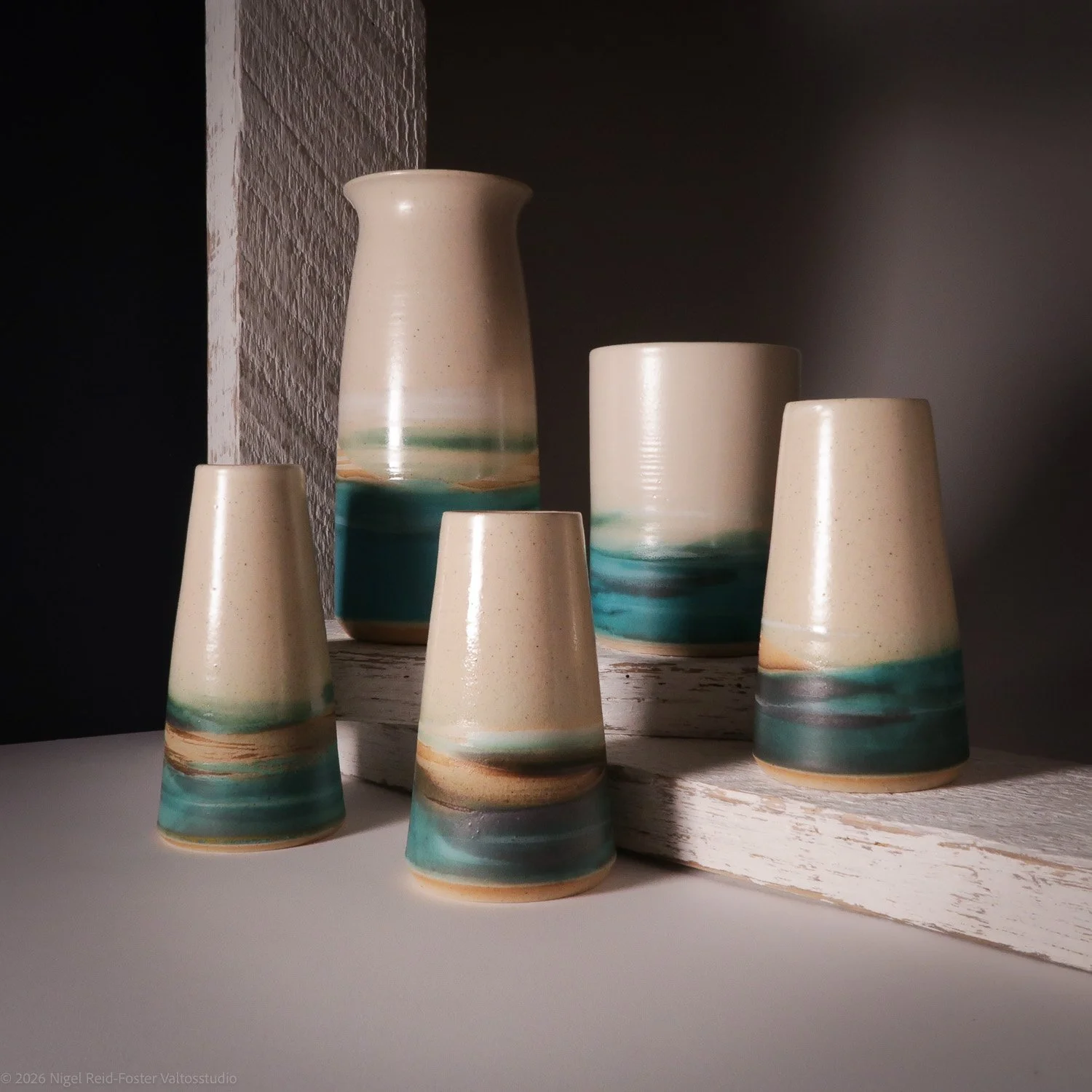 A set of five ceramic vases and cups with a cream-colored base and blue and brown glaze accents, arranged on a white surface and a white wooden platform with a textured gray wall background.