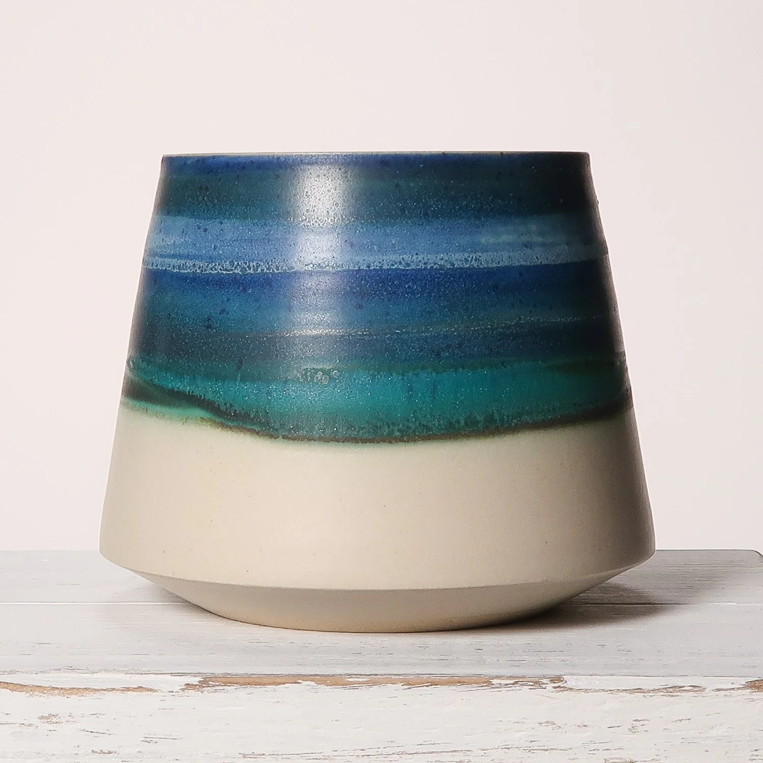 Ceramic vase with a blue and green ombre glaze, placed on a white wooden surface against a plain white background.