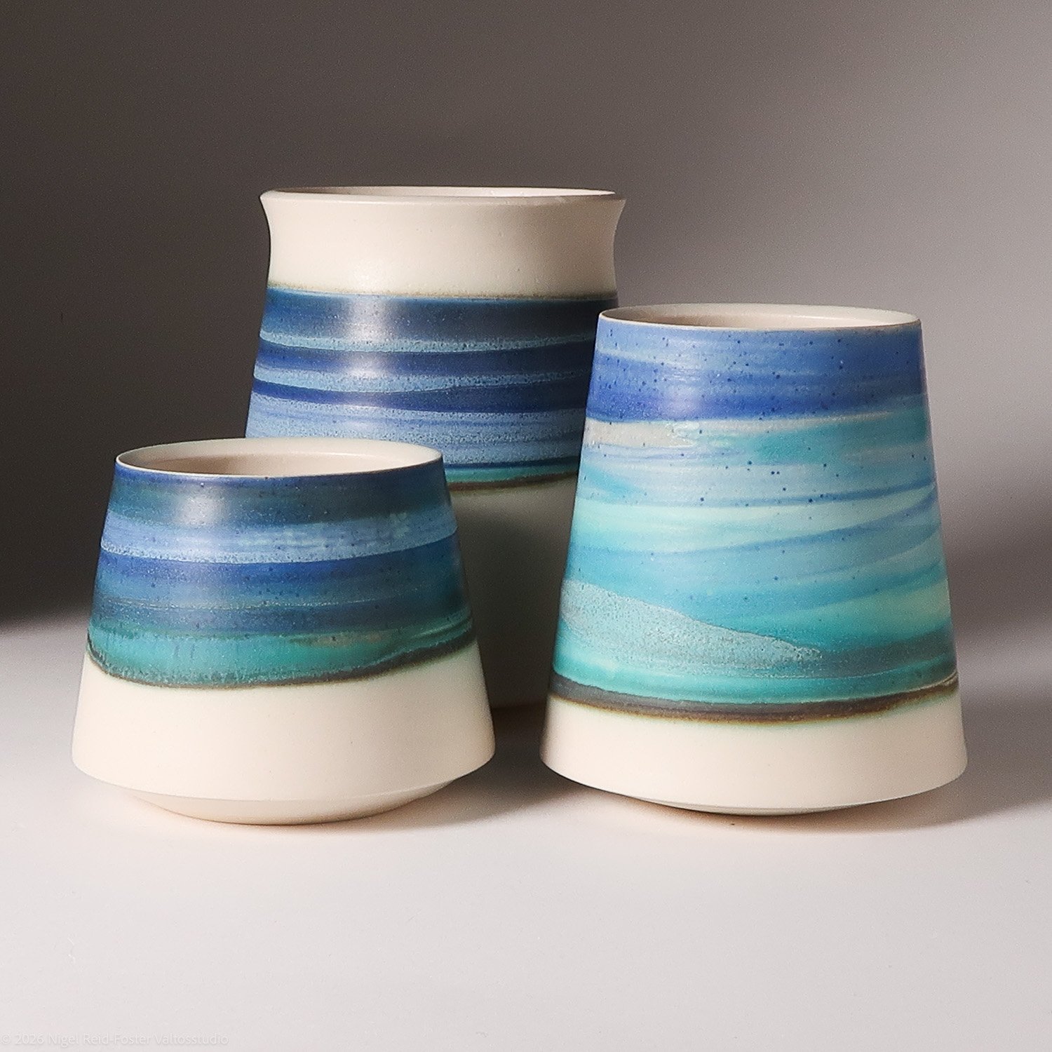 Three ceramic vases with blue, turquoise, and white horizontal striped glaze designs, arranged against a neutral background.