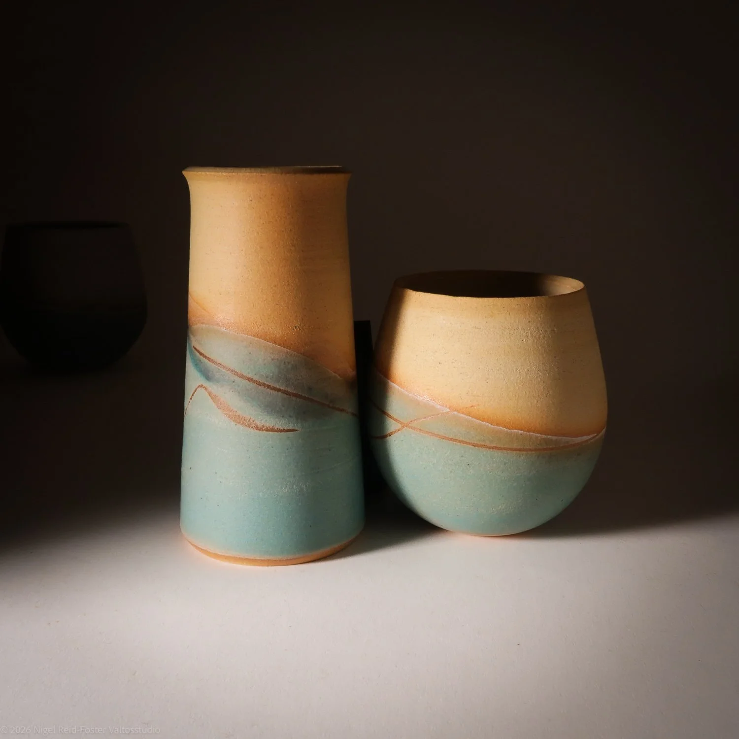 Two handcrafted ceramic vases with light beige and pale blue colors, featuring a subtle wavy design, illuminated by a focused light creating dramatic shadows.