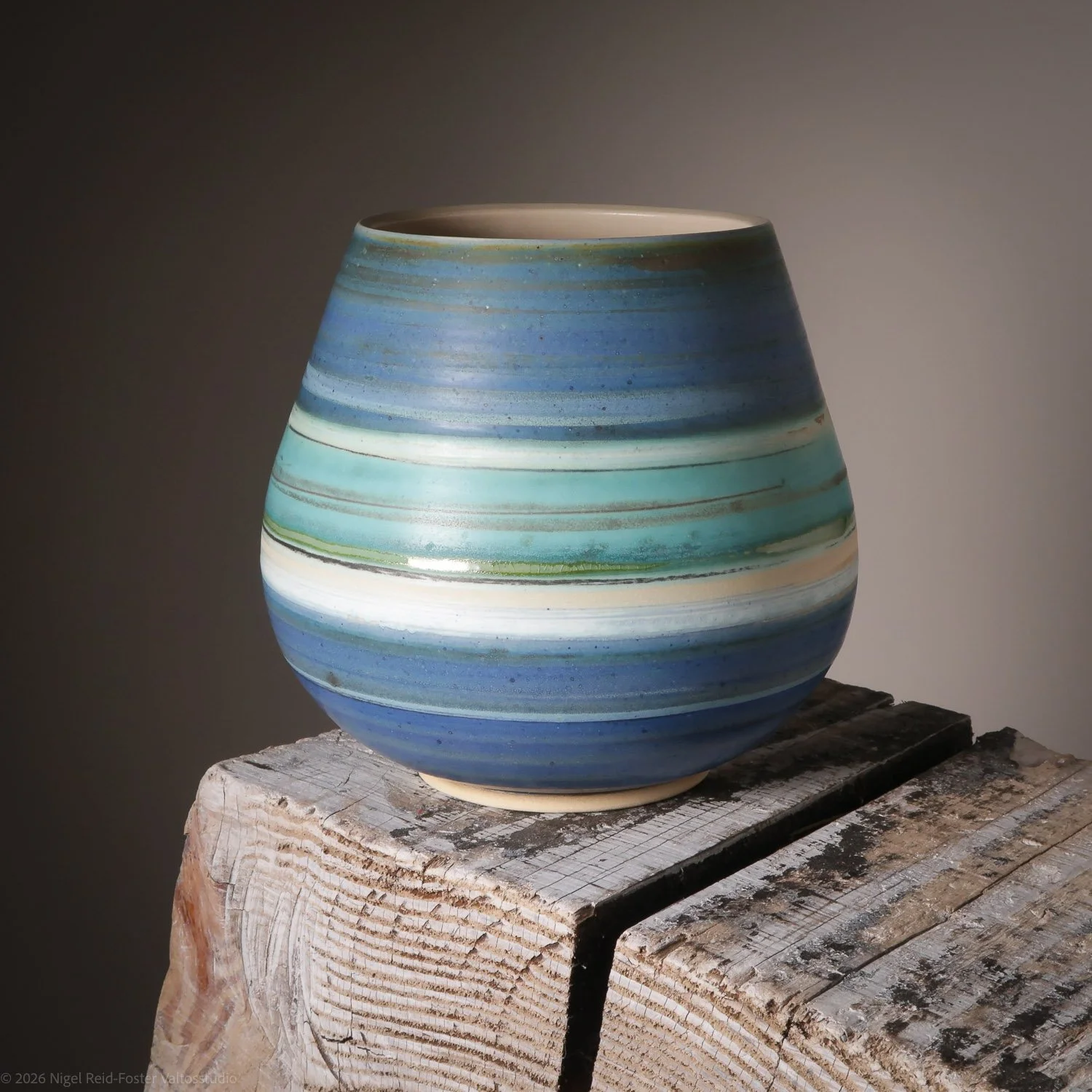 A ceramic vase with blue, green, and white horizontal stripes sits on a weathered wooden block against a neutral background.