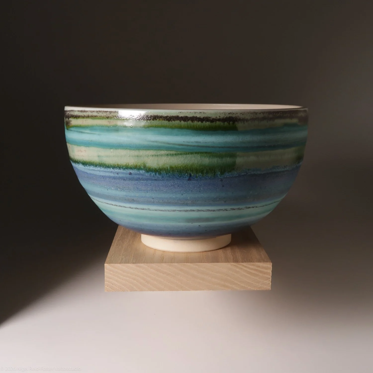 A colorful, hand-painted ceramic bowl with blue, green, and black stripes, placed on a small wooden stand against a dark background.