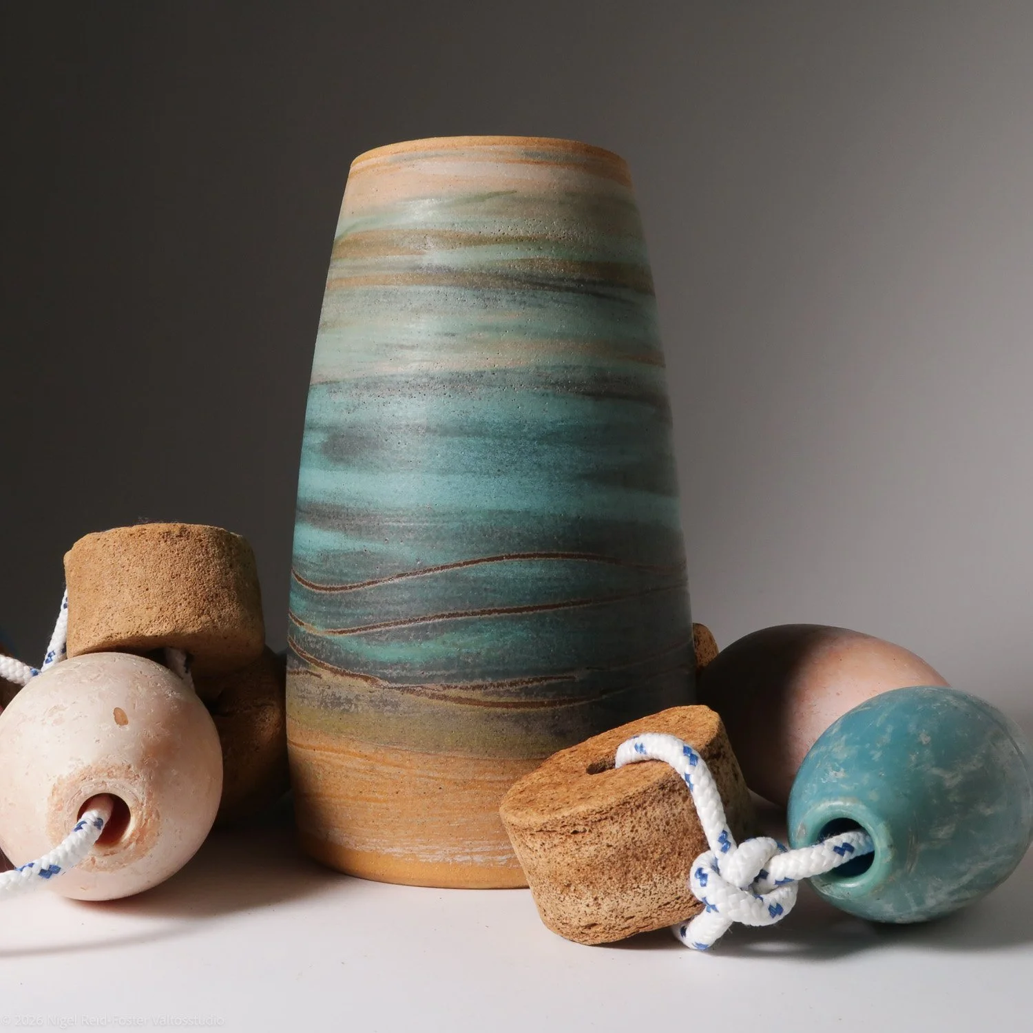A large pottery vase with a marbled blue, green, and beige pattern, surrounded by smaller ceramic and foam buoys with strings, on a plain surface against a plain gray background.