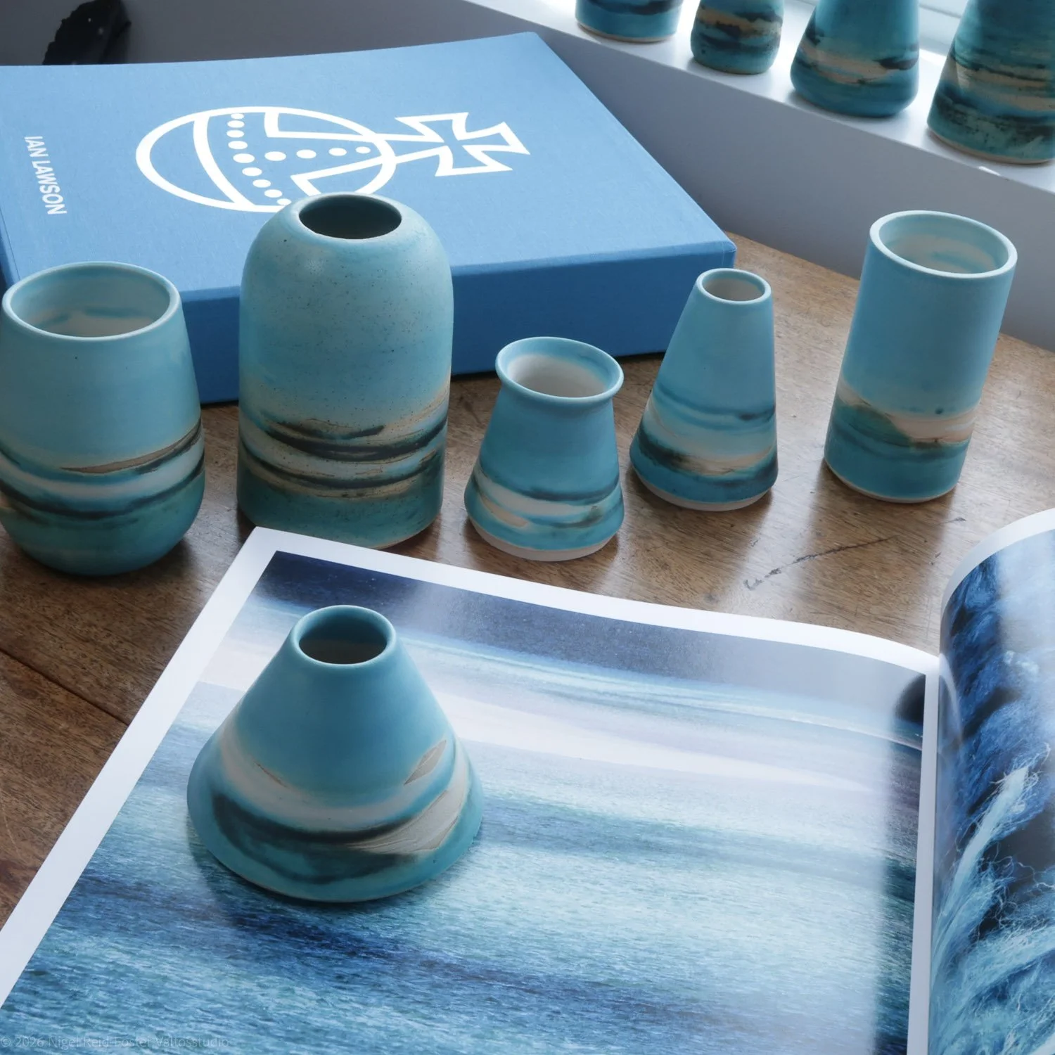 Set of five ceramic vases in various sizes with blue and white marbled glaze, arranged on a wooden surface. A blue box with a white logo and the name 'Ian Lawson' is in the background, and an open book with a blue ocean scene is in the foreground.